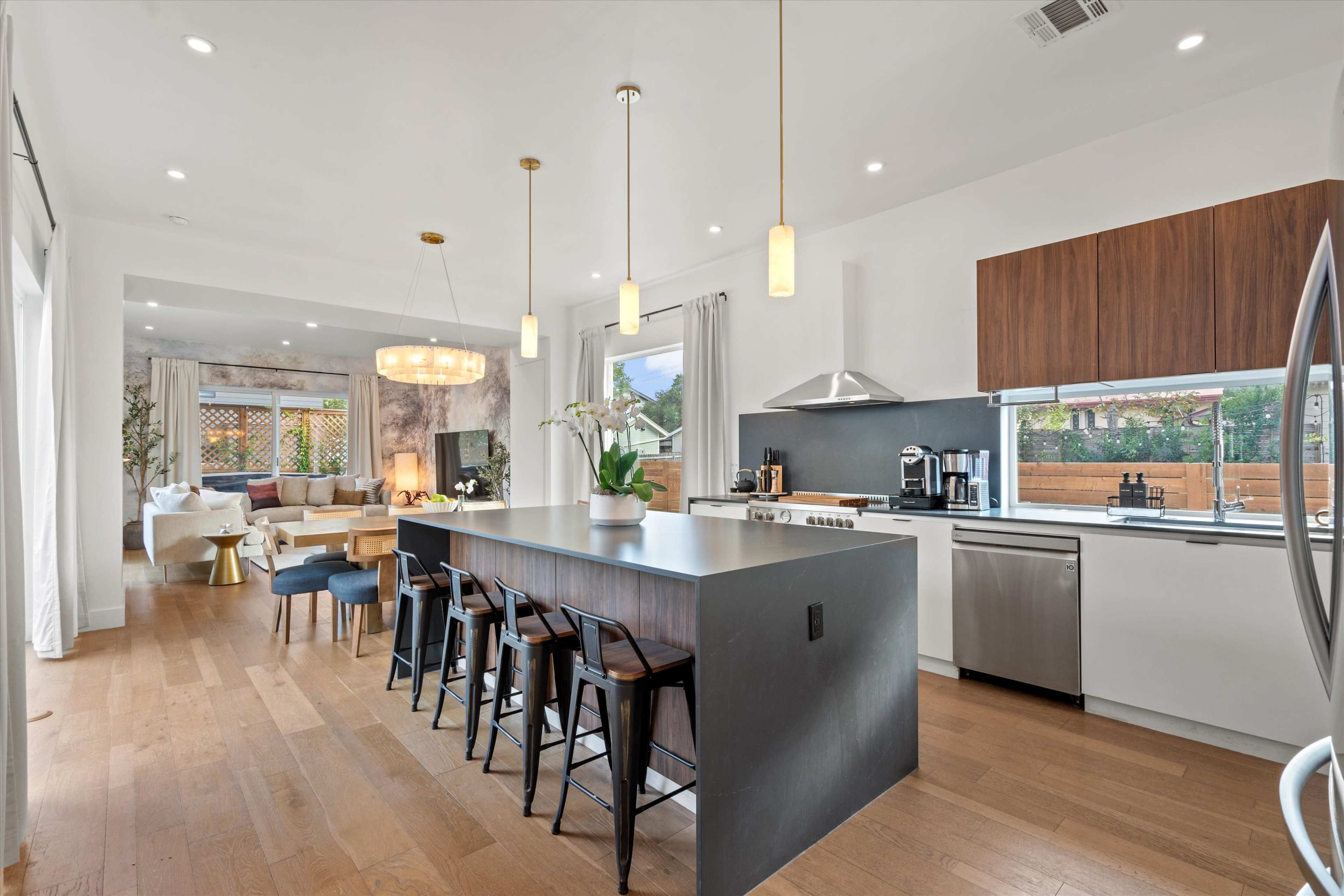 The image shows a modern kitchen with a large island and seating, connected to a bright living area featuring a sofa and decorative lighting.