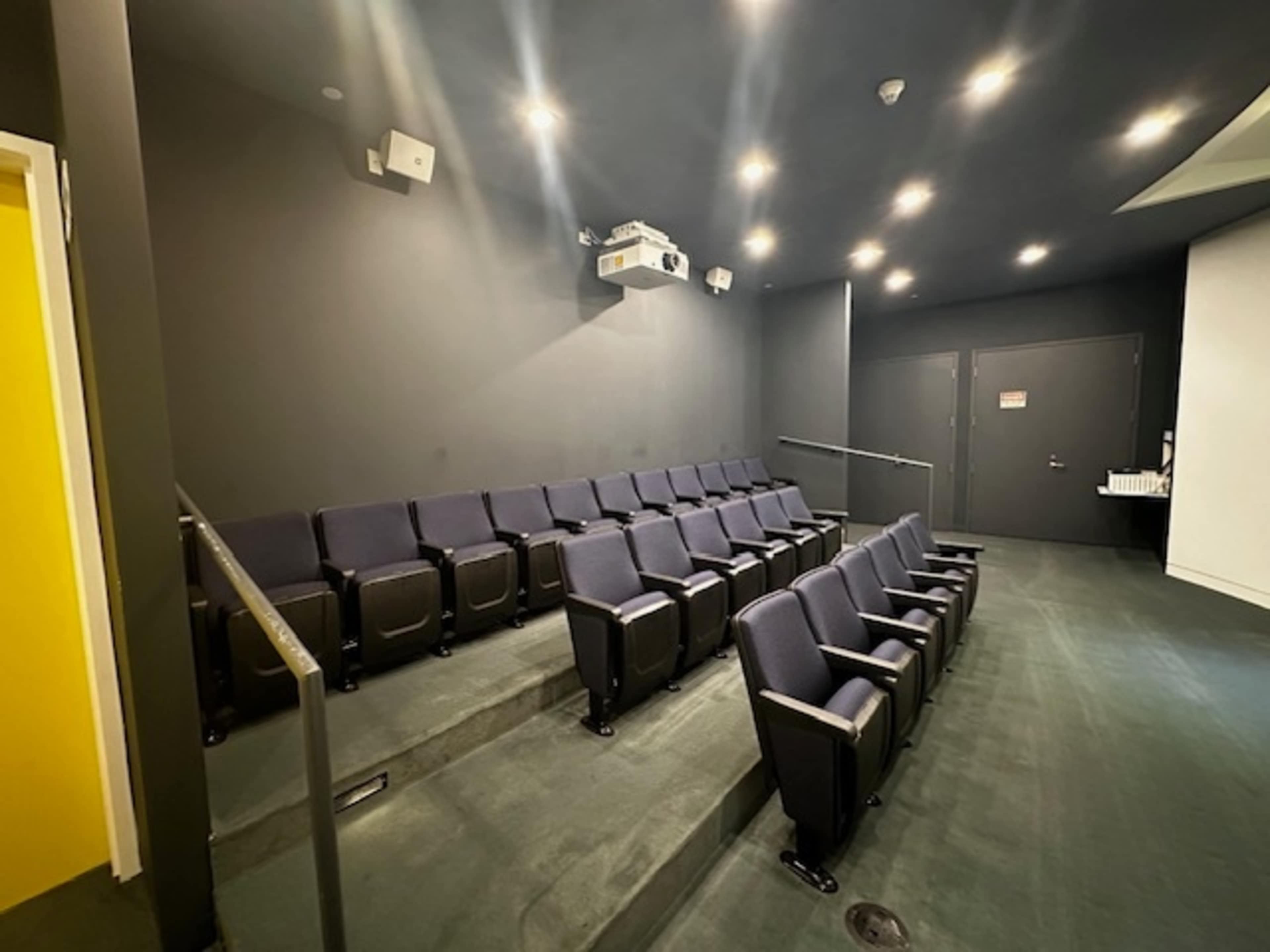 A small auditorium with several dark blue seats arranged in tiers facing a projection screen.