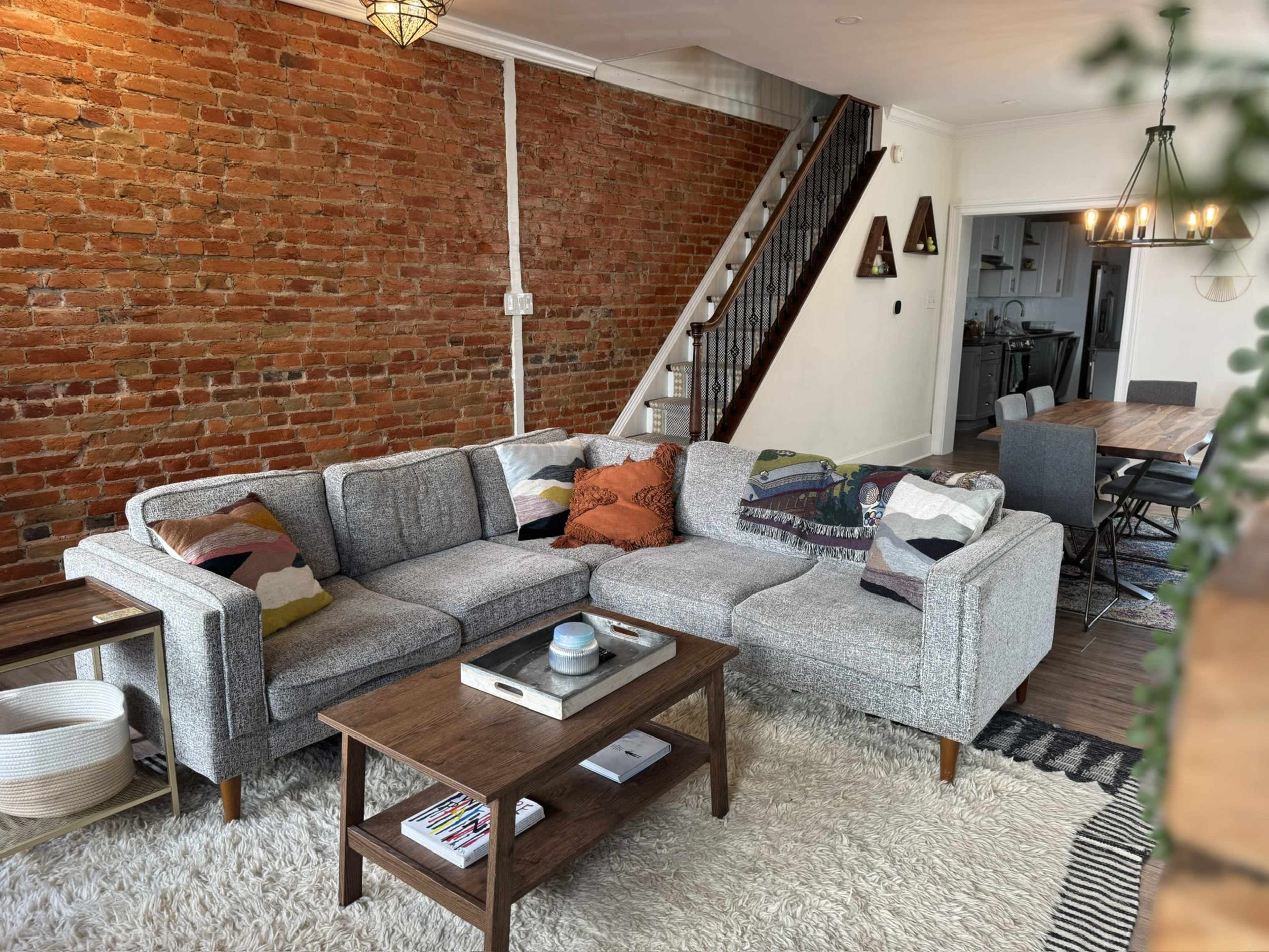 The image shows a living room with a gray sectional couch, a wooden coffee table, and a visible brick wall, along with a dining area and staircase in the background.