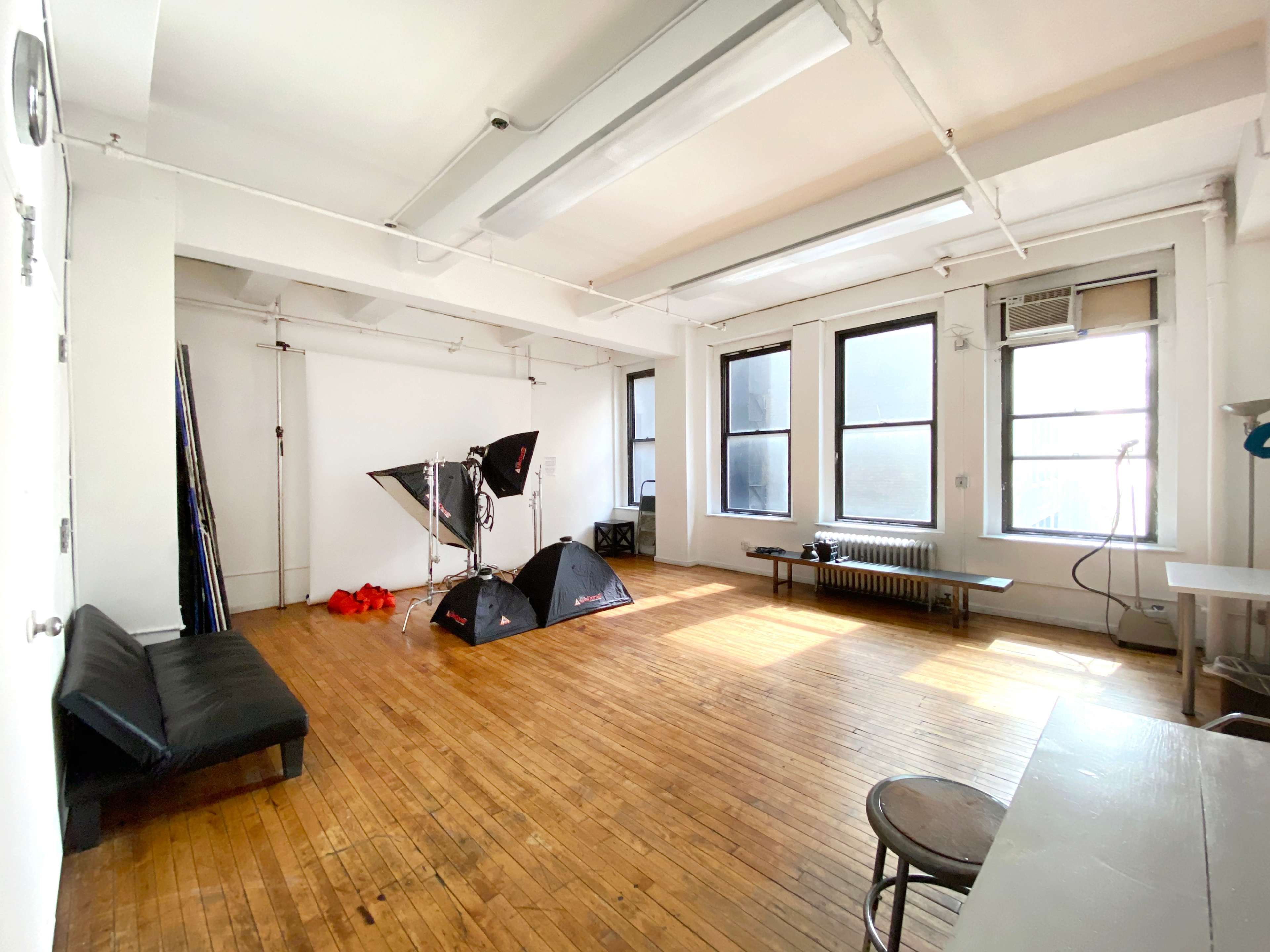 A spacious, well-lit studio features wooden floors, large windows, and photography equipment set up in the center.