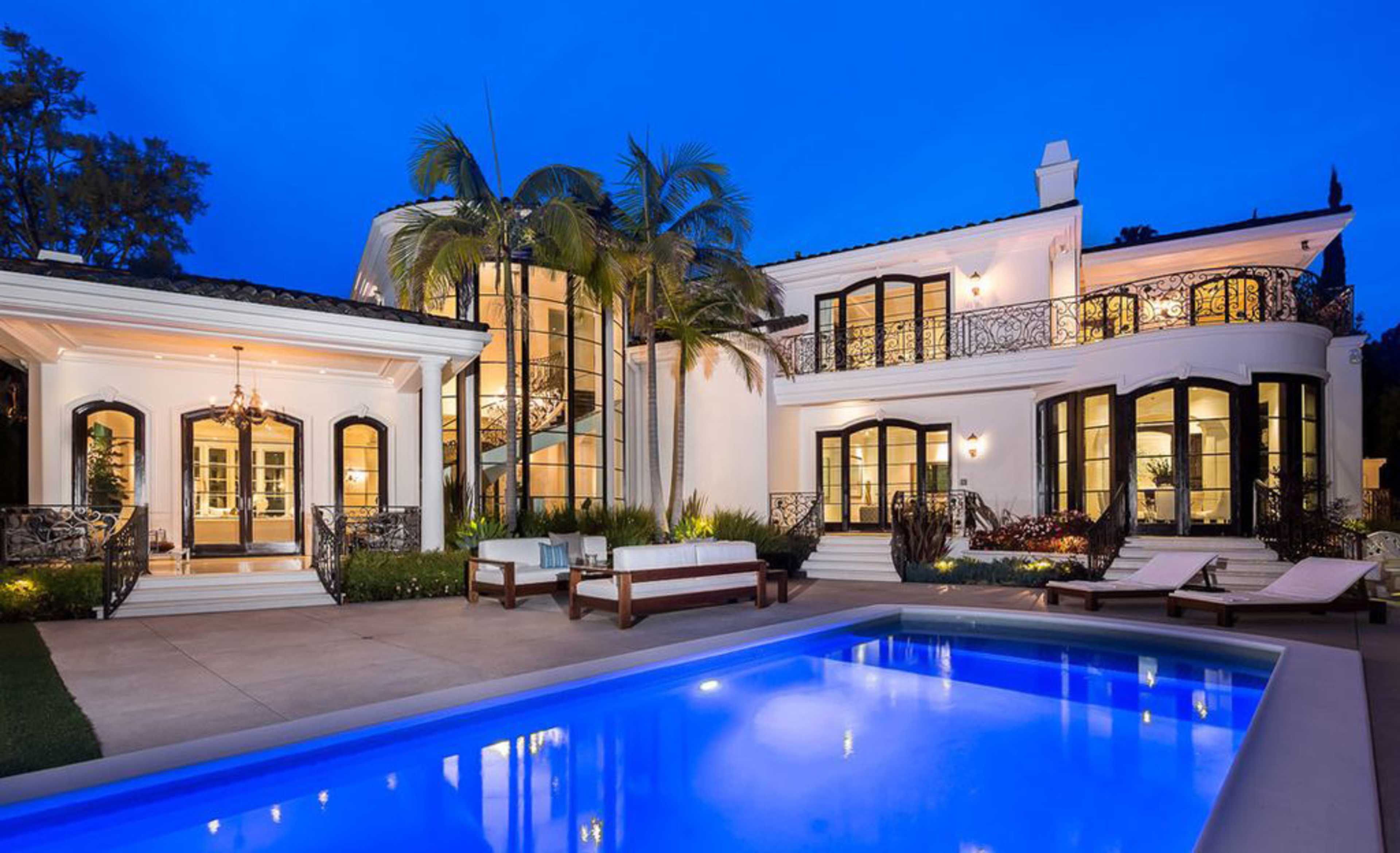 A modern two-story house with large windows, a swimming pool in the foreground, and lush landscaping under a twilight sky.
