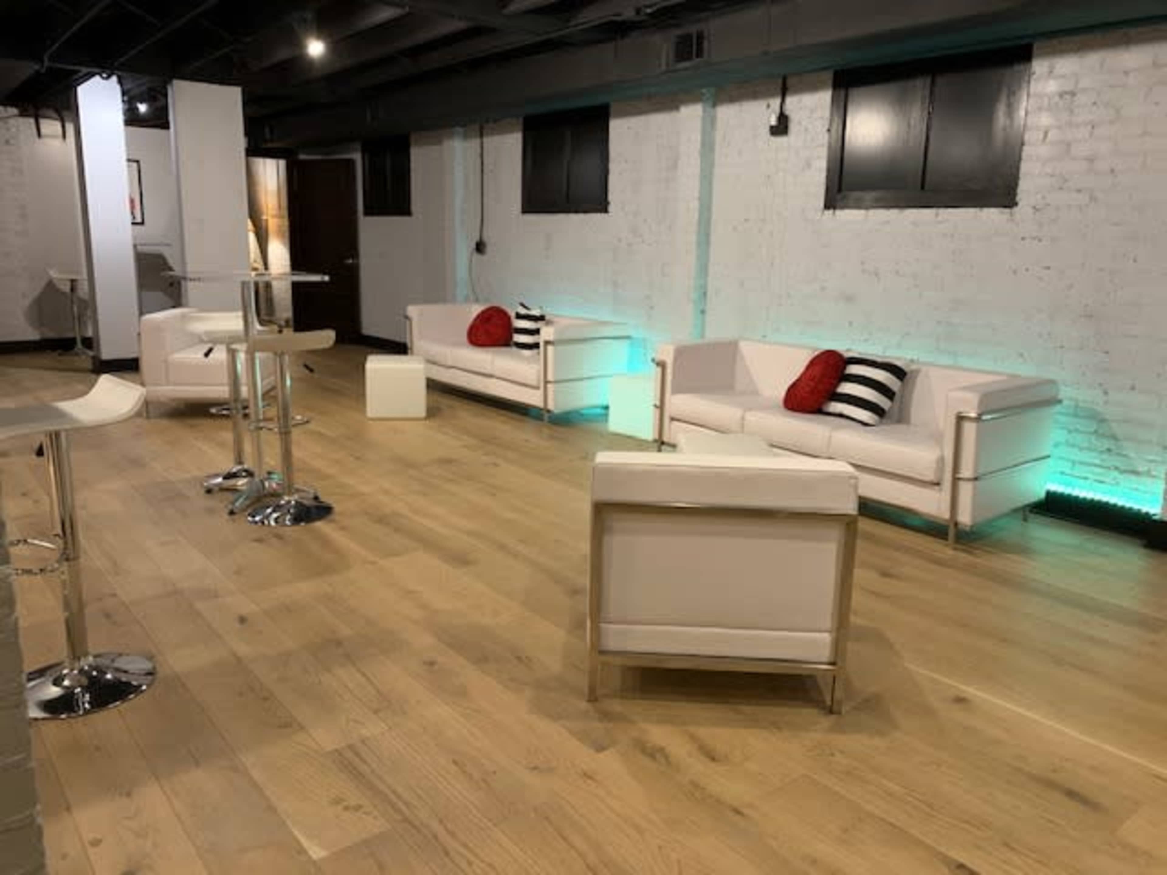 A modern, spacious room with white brick walls, wooden flooring, and minimalist white furniture, including sofas and tables, illuminated by ambient lighting.