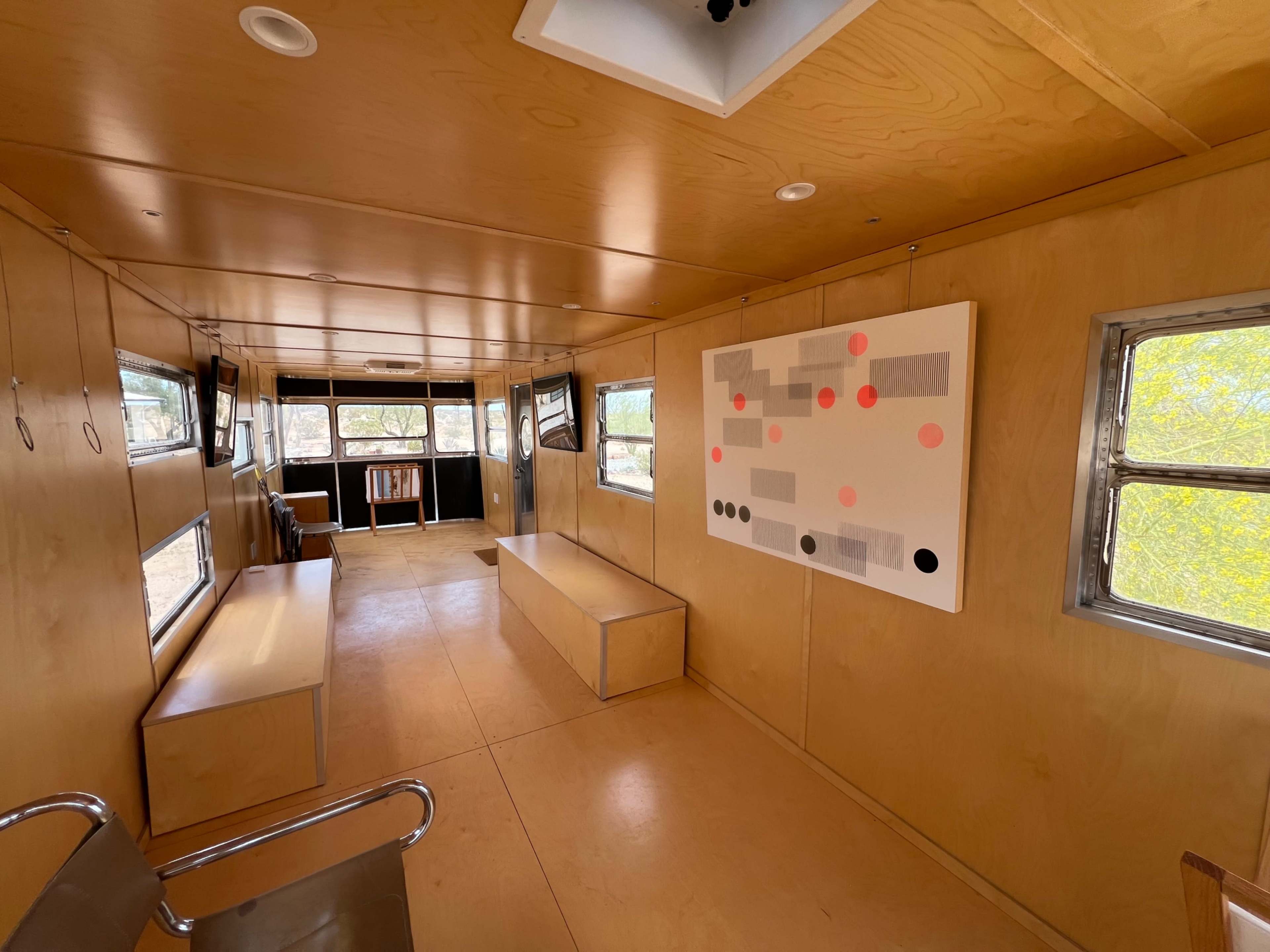 The image shows the interior of a minimalist cabin with wooden walls, large windows, and a simple layout featuring a central bench and an abstract artwork on the wall.