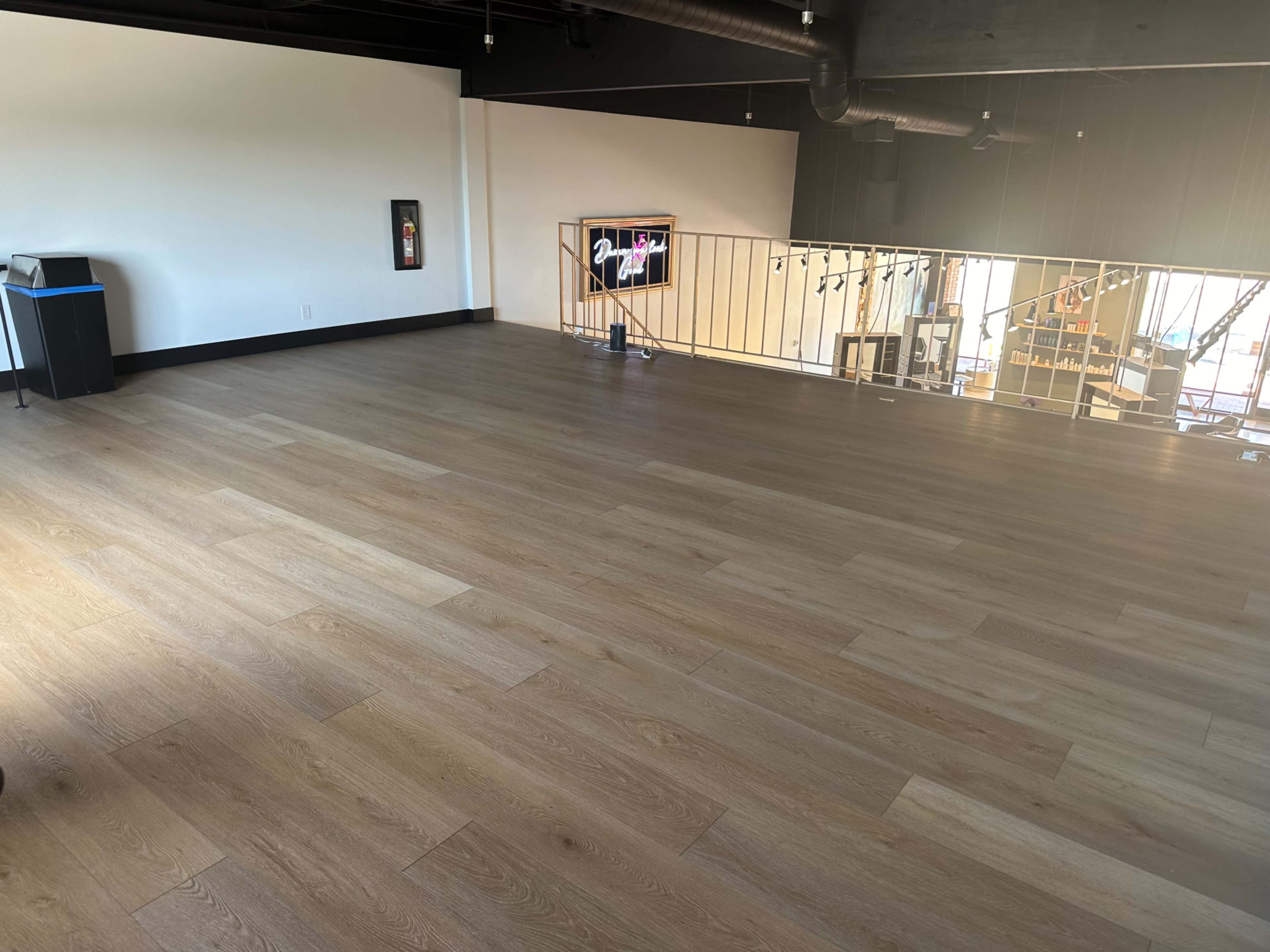 The image shows an empty, well-lit room with wooden flooring and large windows along one wall.
