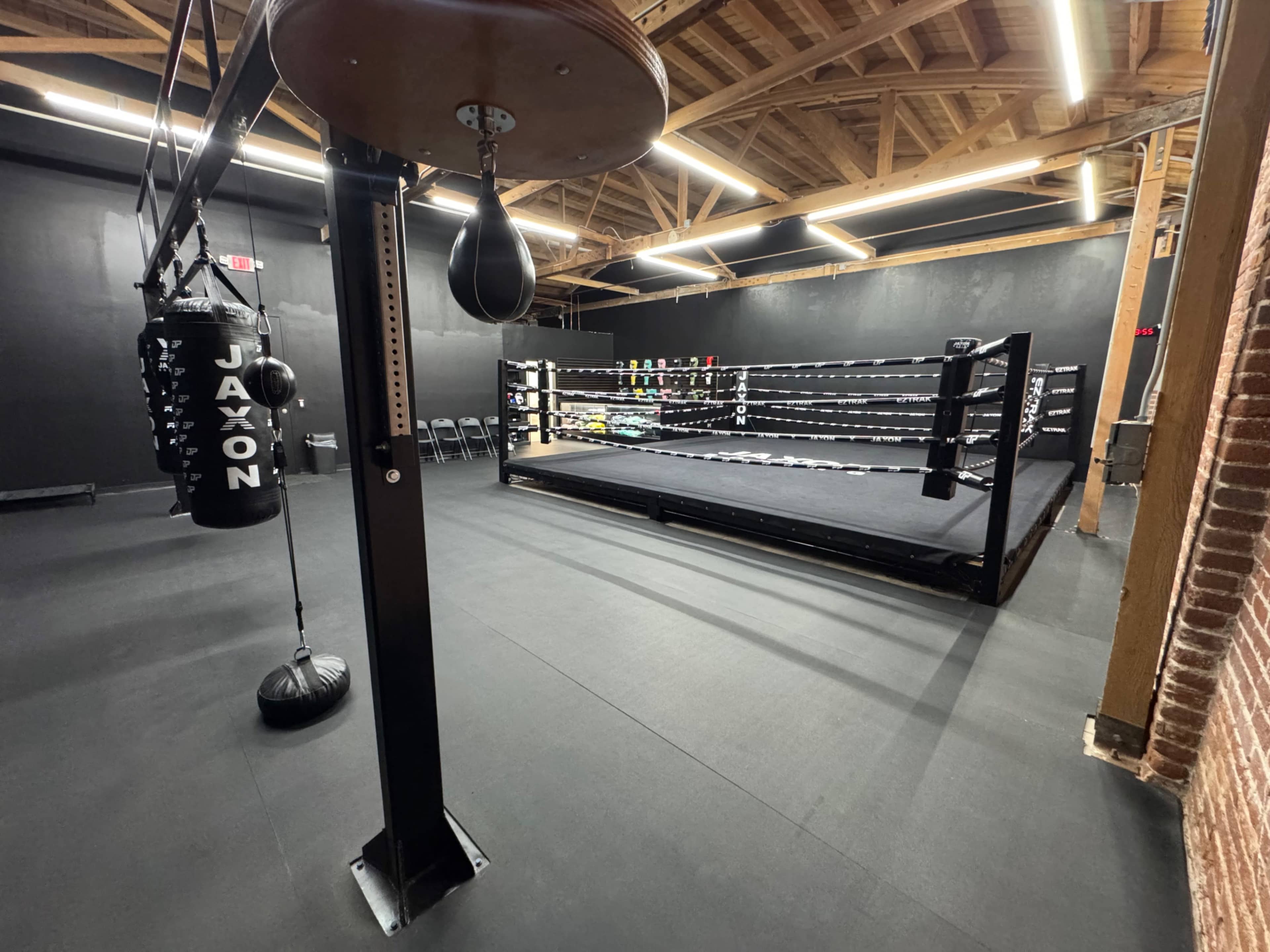 The image shows a boxing gym featuring a boxing ring, heavy bag, and training equipment against a backdrop of black walls and wooden beams.