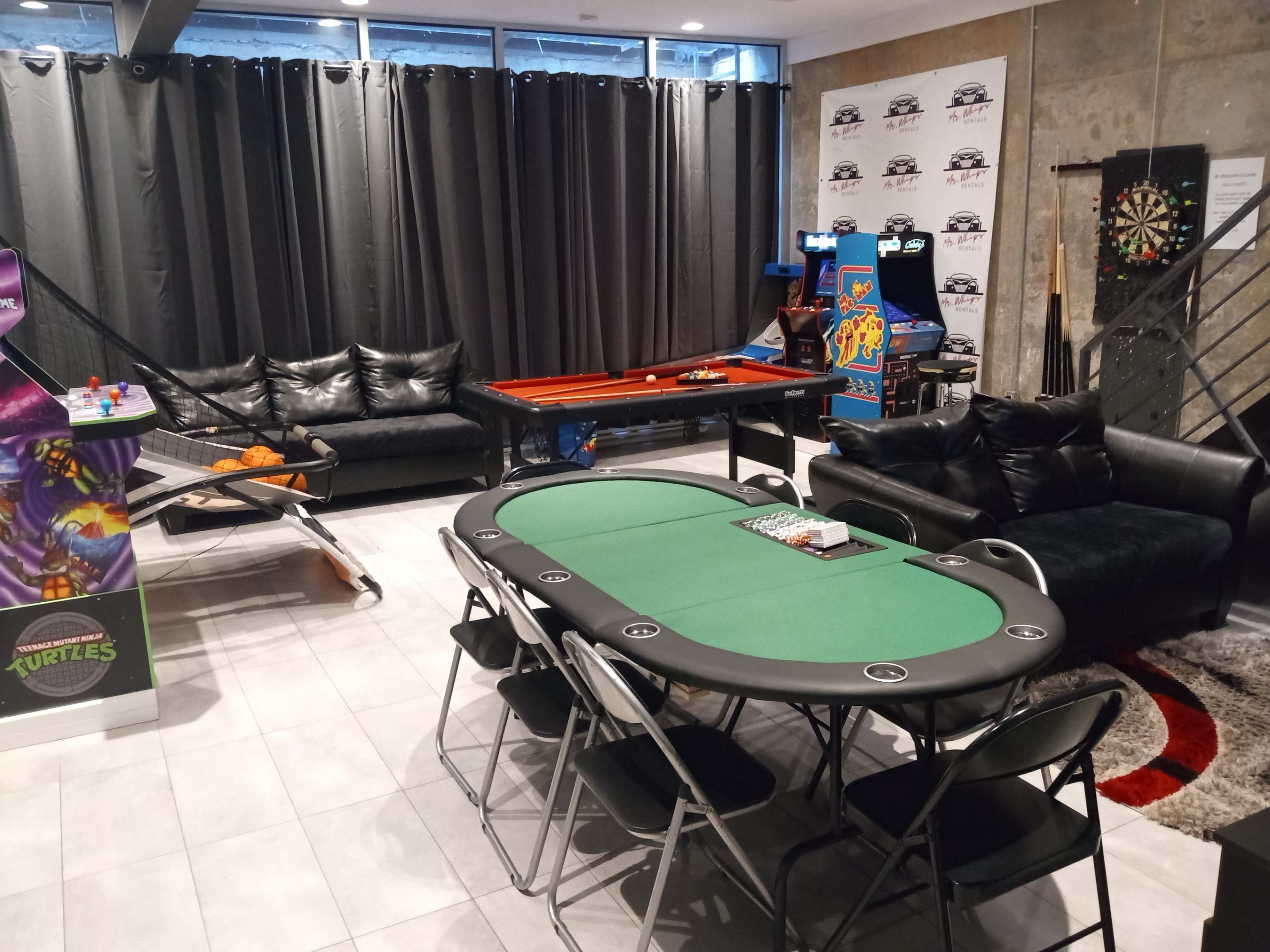 The image shows a recreation room featuring a pool table, a green poker table surrounded by black chairs, a dartboard, and various arcade games, with seating areas in the background.