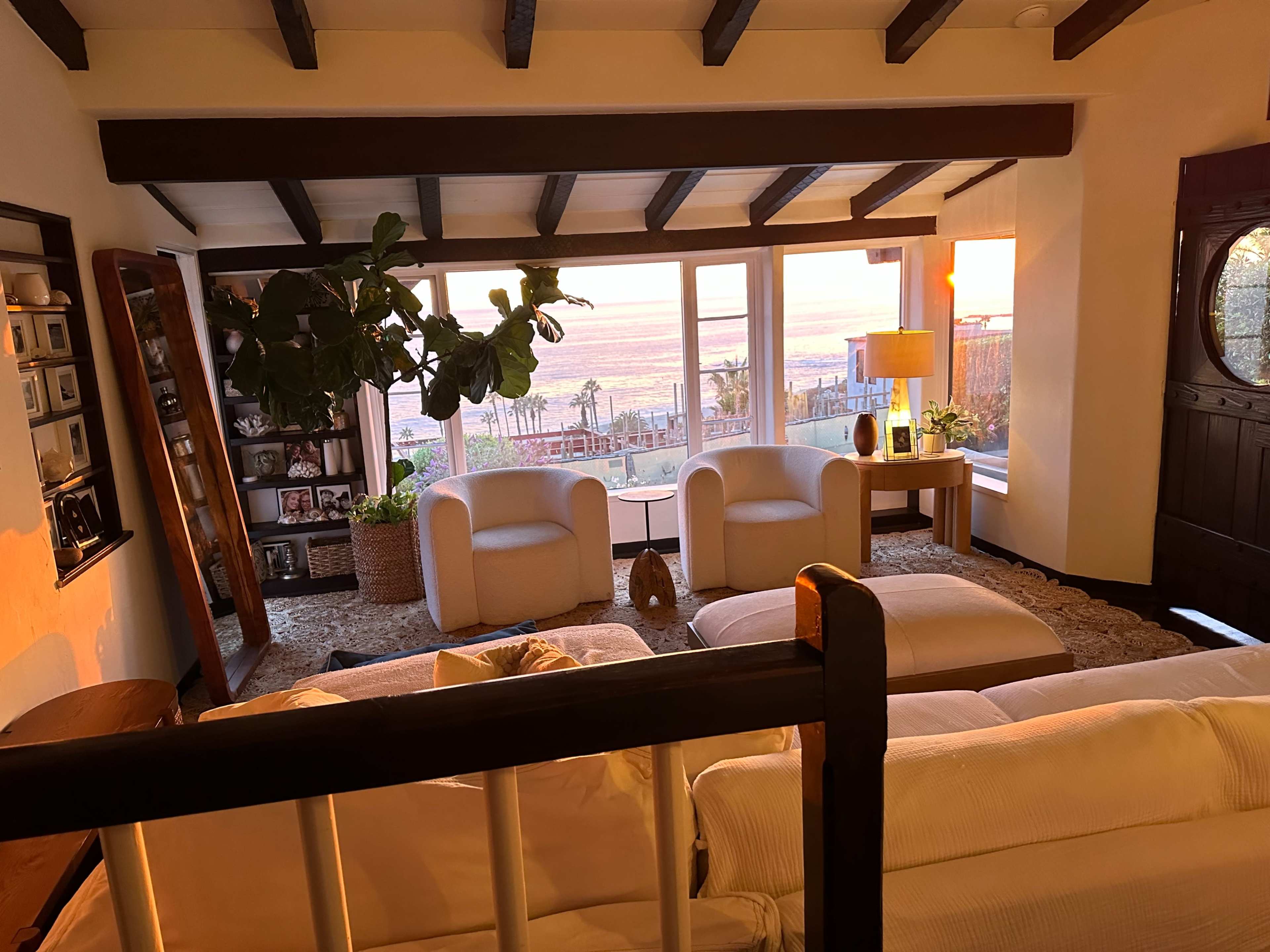 A bright living room with two white chairs, large windows overlooking a sunset, and a potted plant near a lamp.