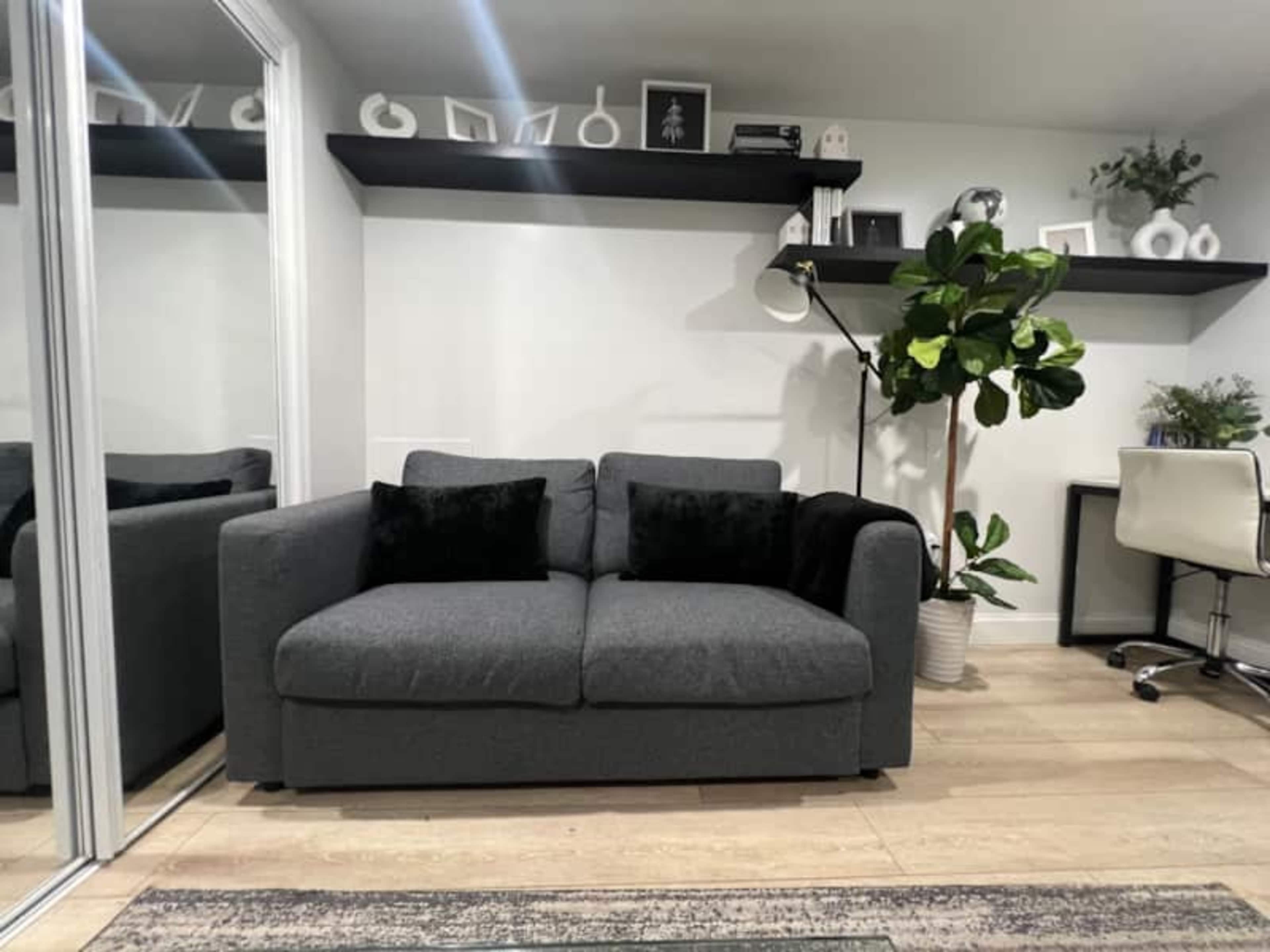 The image shows a modern living space featuring a gray sofa with black throw pillows, a potted plant, and a desk with a chair, all set against a light wall and mirrored closet.