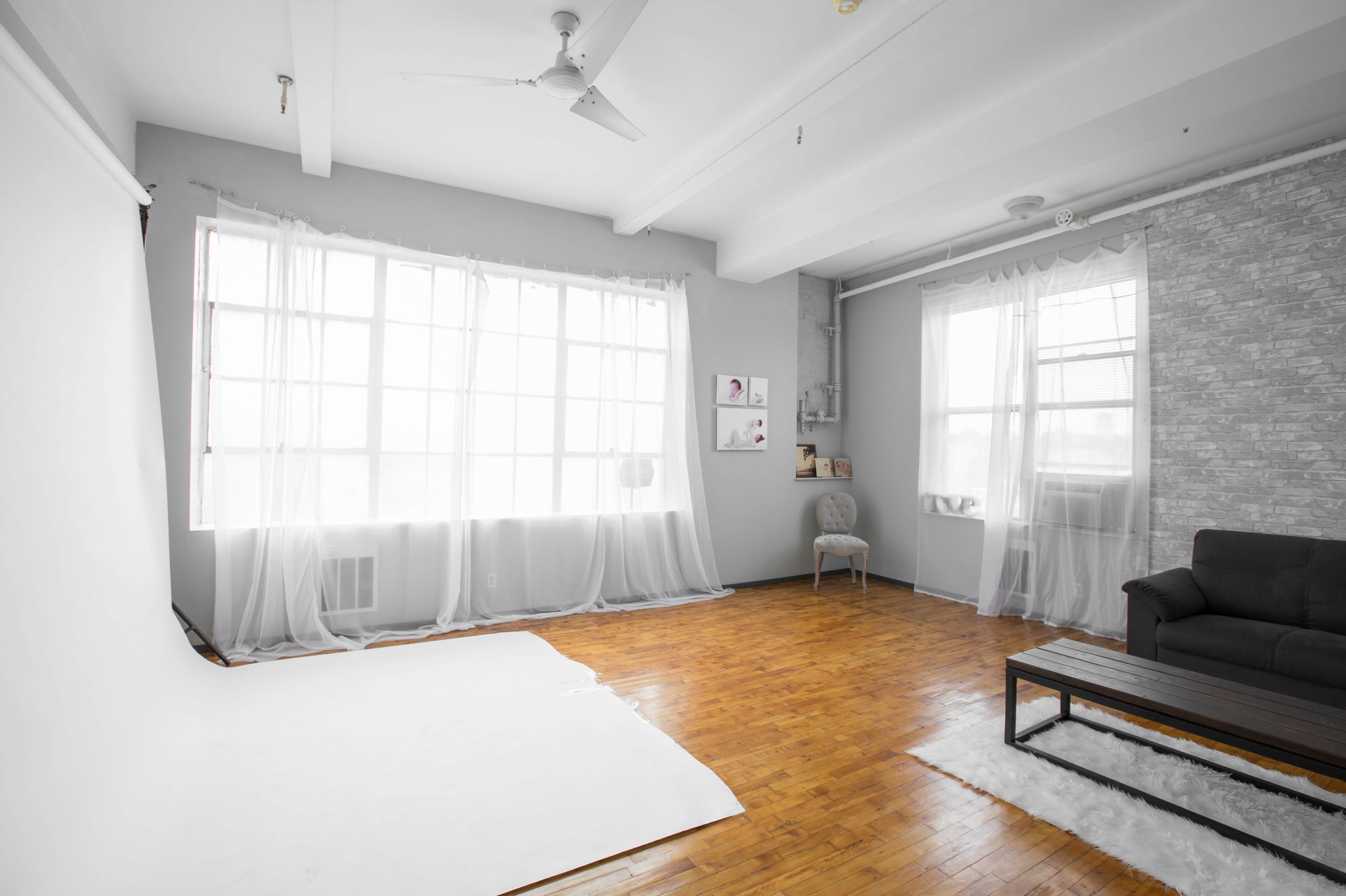 The image shows a simple, bright room with wooden flooring, large windows covered by sheer curtains, and minimal furnishings including a dark sofa and a coffee table.