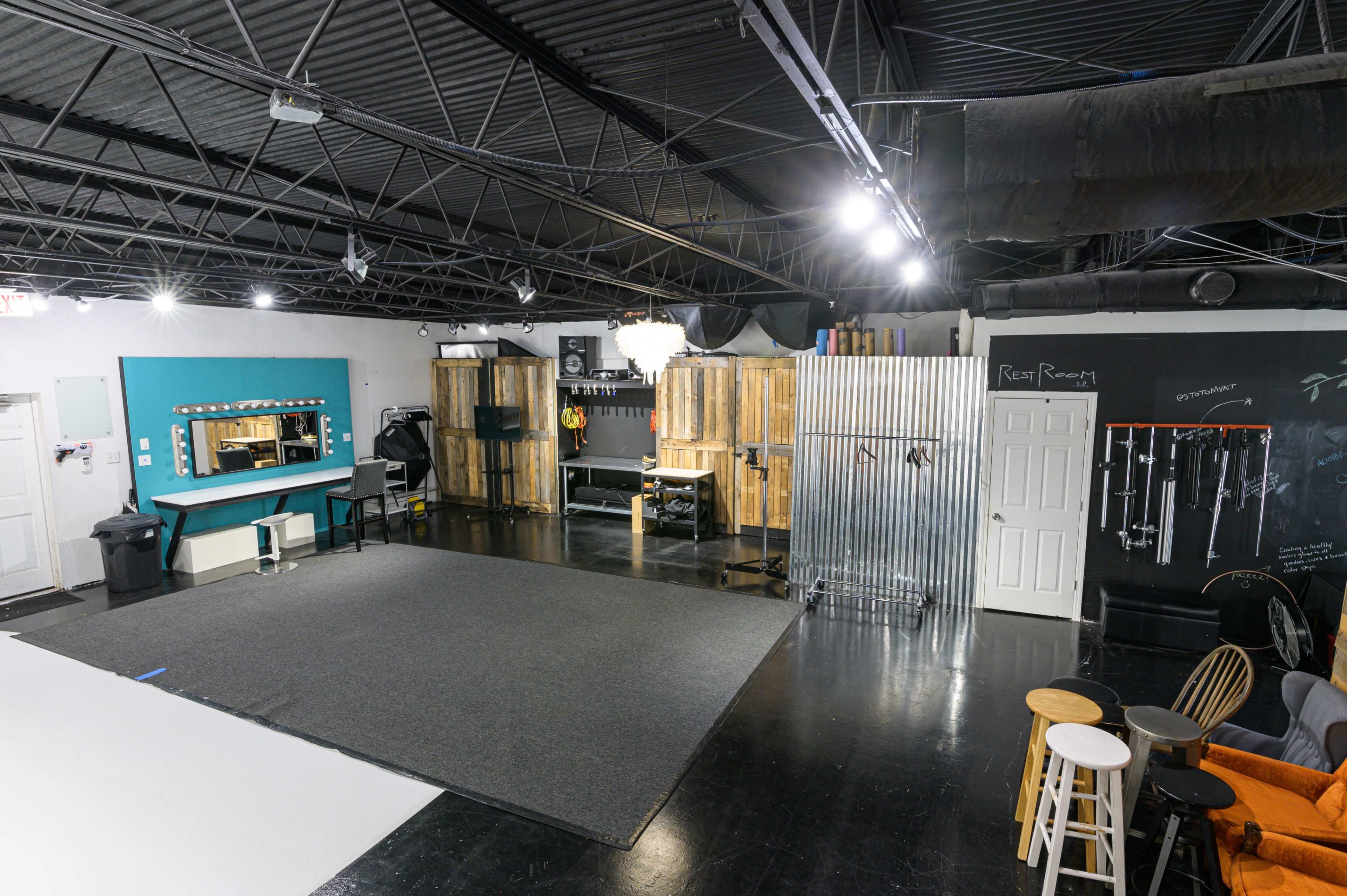 A studio space featuring a large carpeted area, mirrored wall with a blue accent, wooden storage units, and various equipment against a black chalkboard wall.
