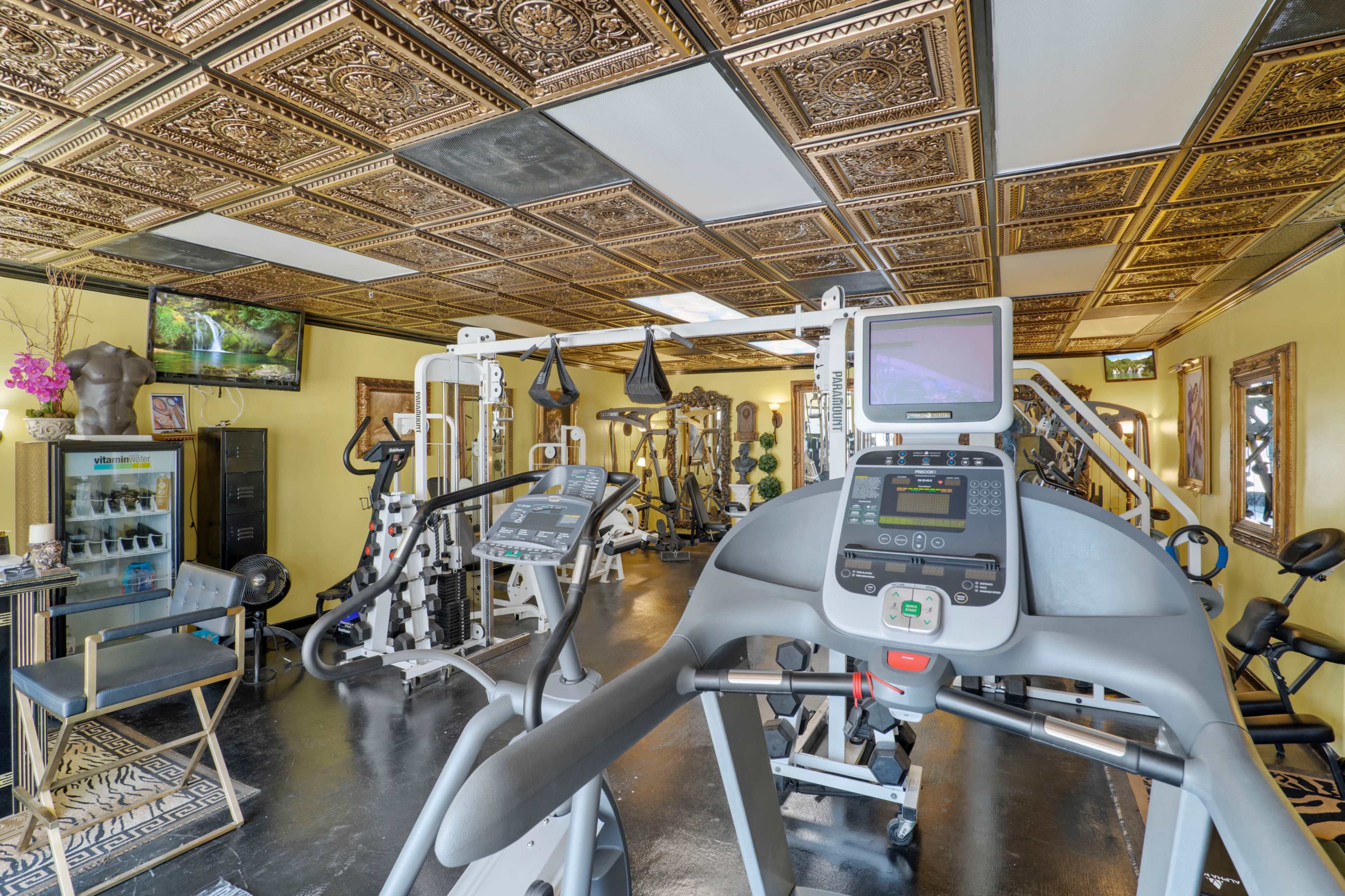 A fitness room with various gym equipment, decorative gold ceiling tiles, and a large mirror on one wall.