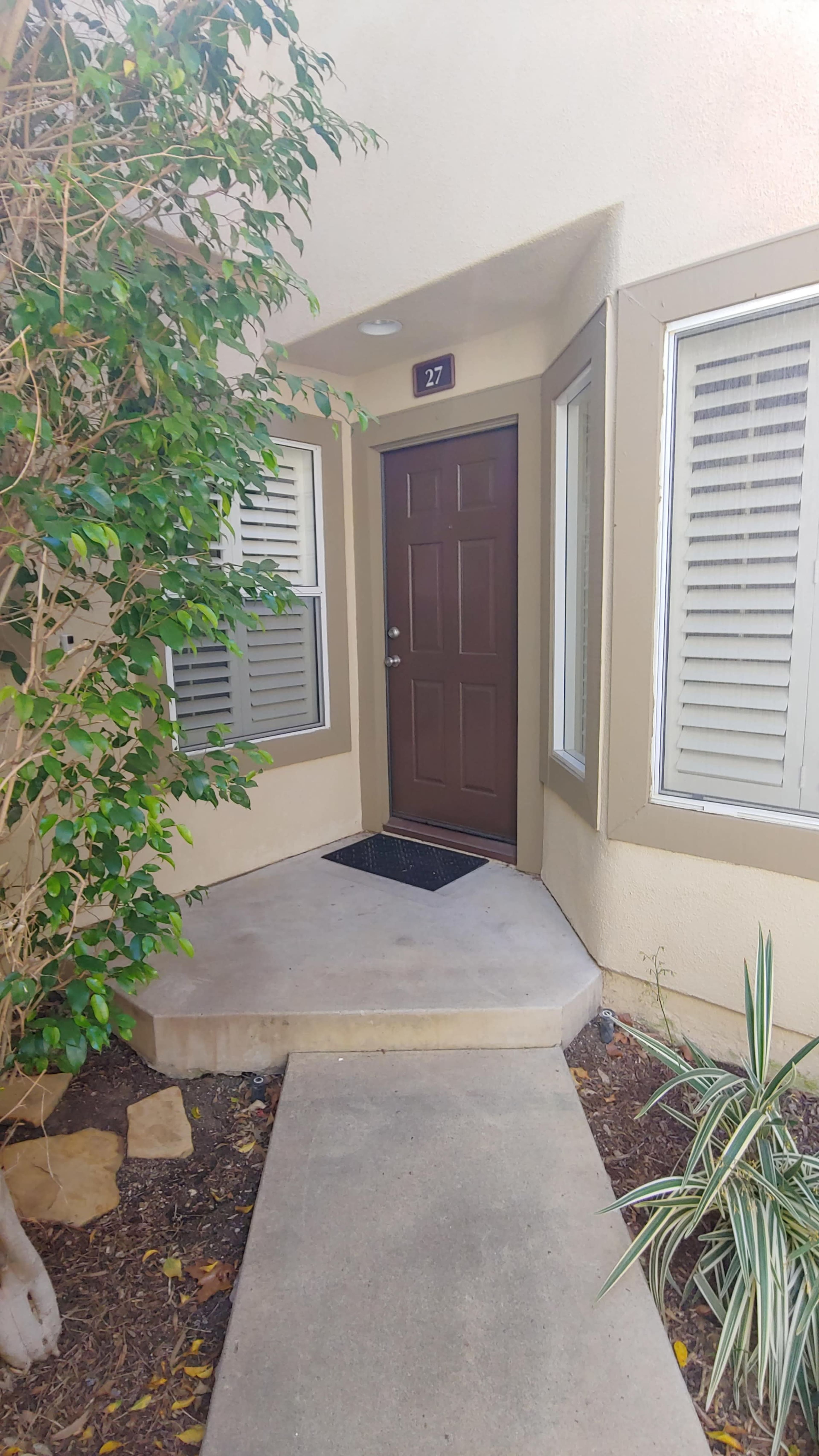 A front porch area with a door numbered 27, surrounded by plants and a walkway leading up to the entrance.