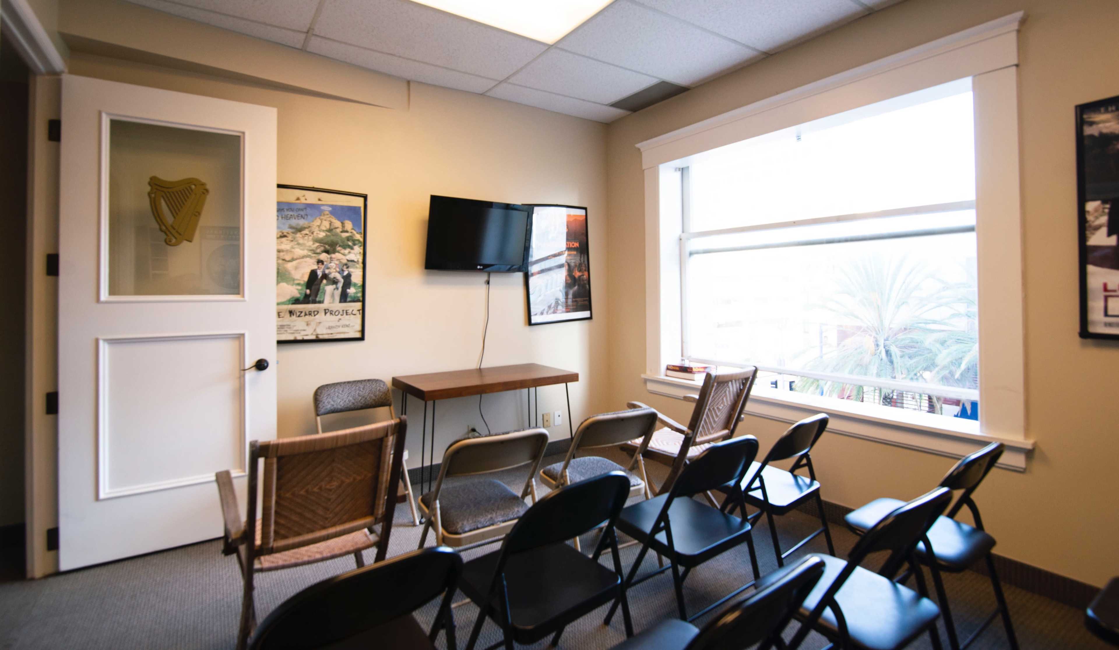 A meeting room features a small table, a television mounted on the wall, and several black folding chairs arranged in rows.