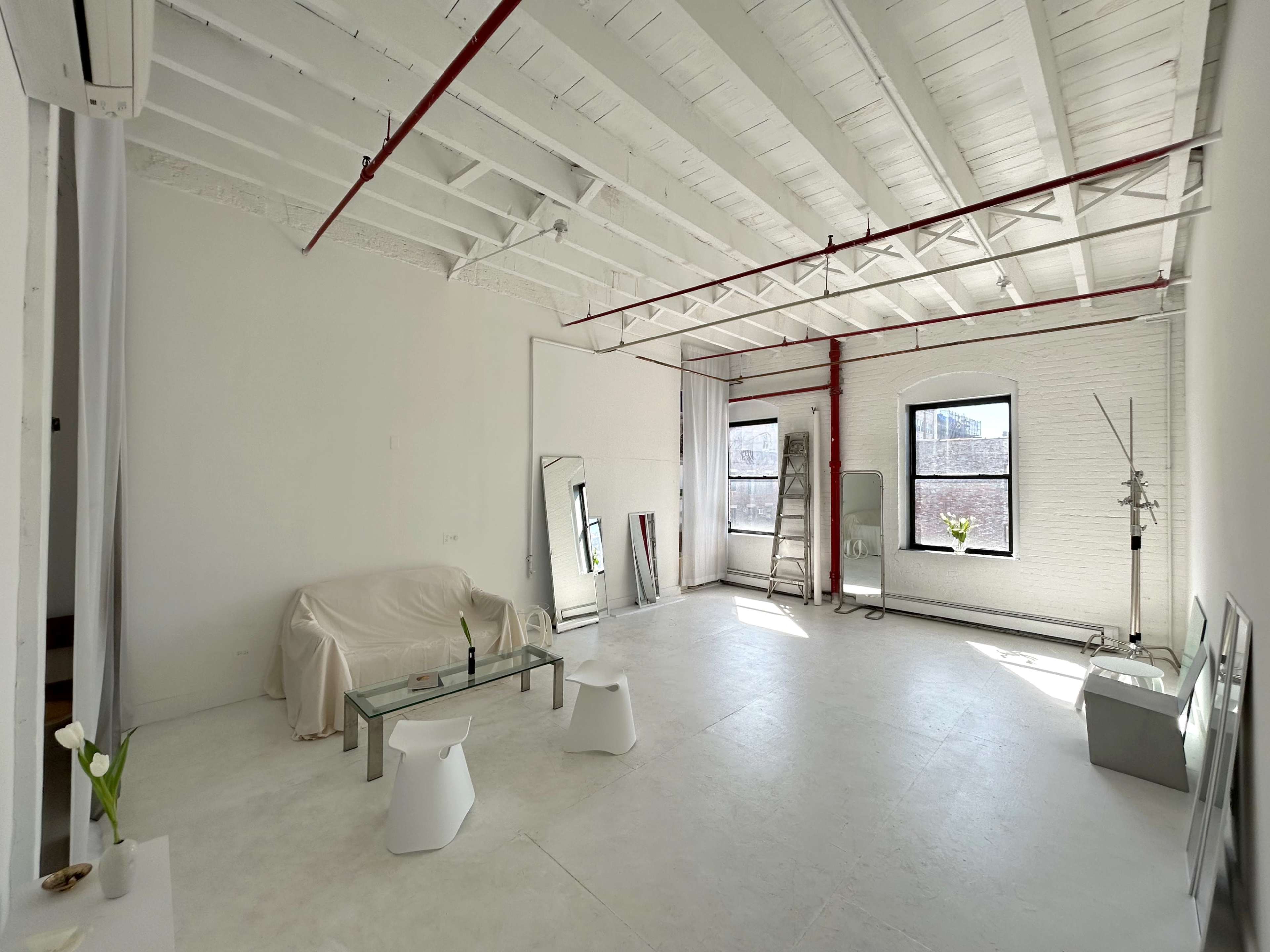 A spacious, empty studio with white walls and a high ceiling, featuring a sofa covered with a sheet, a glass coffee table, and minimal furnishings.