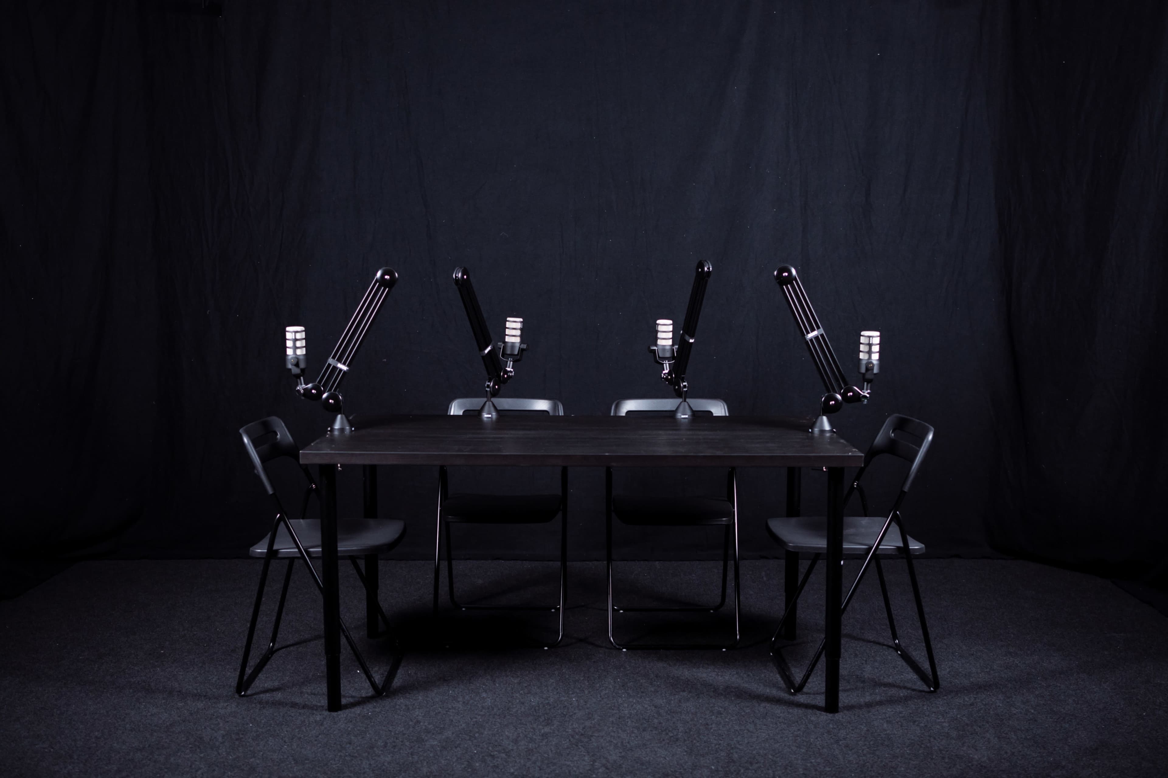 A black table with four chairs and mounted microphones is set against a dark backdrop.