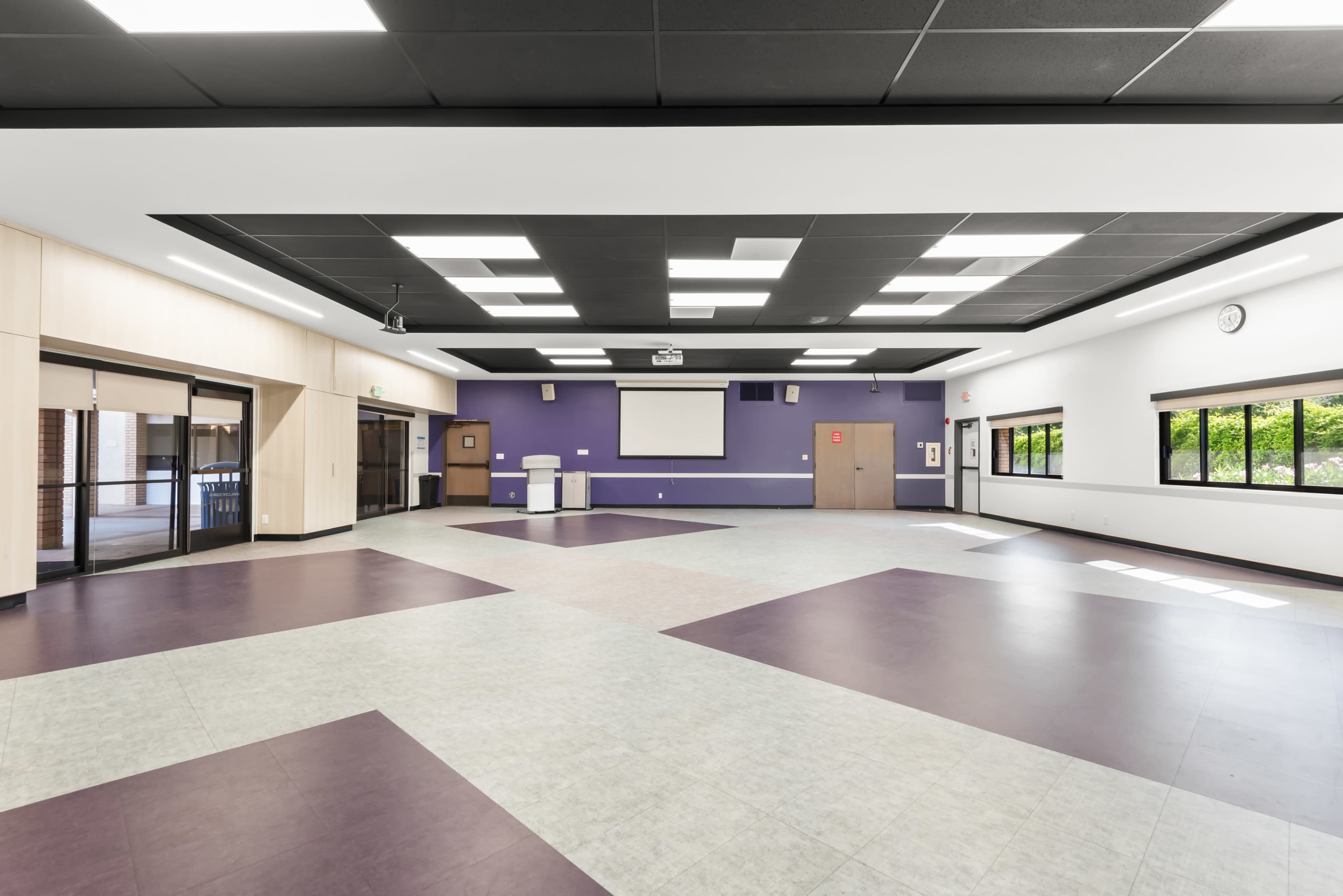 The image shows a large, empty multi-purpose room with a mixture of light and dark flooring, large windows, and a projector screen on the wall.