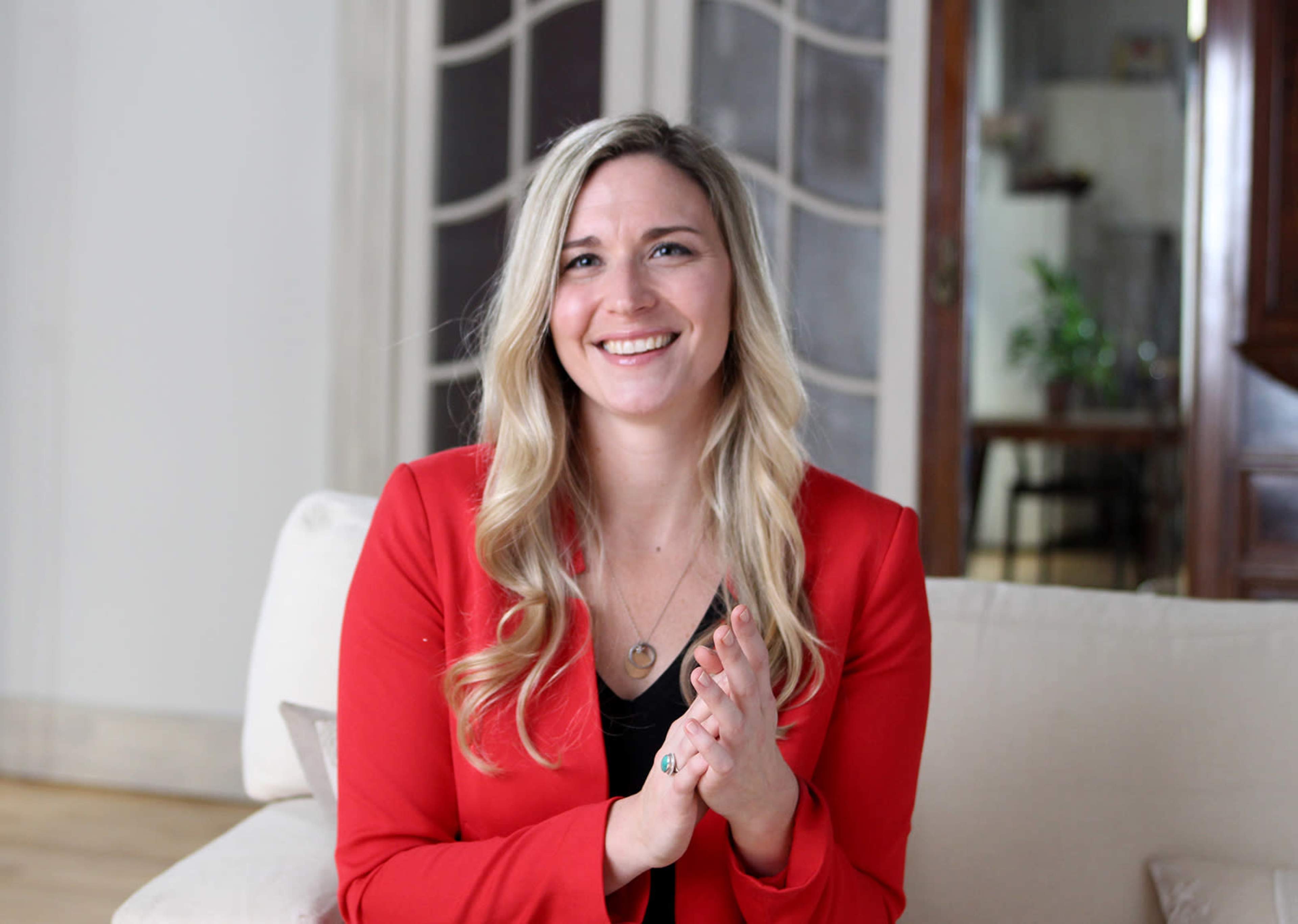 A smiling woman with long, wavy hair in a red blazer is clapping her hands while sitting on a couch in a well-lit room.