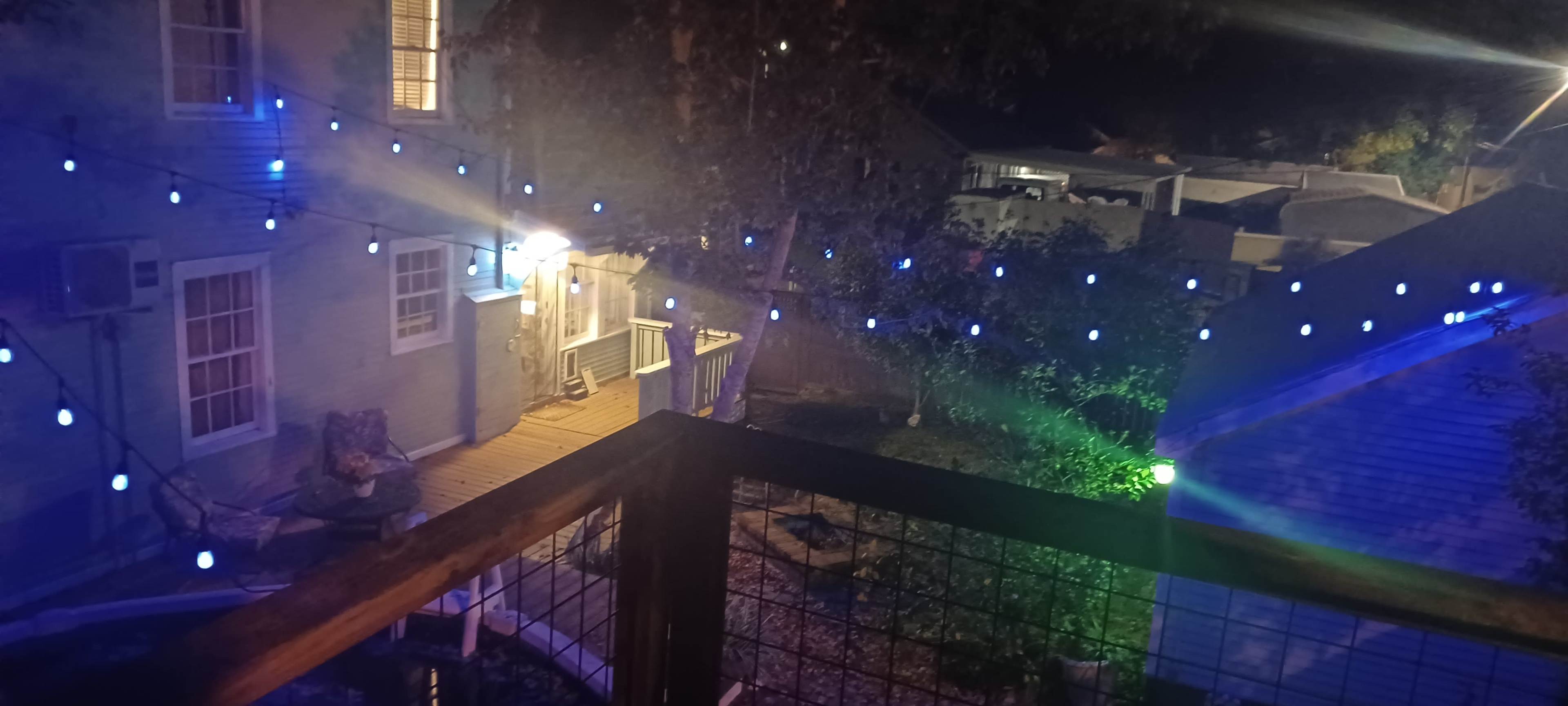 The image shows a backyard scene at night, with blue string lights illuminated above a wooden deck and a house in the background.