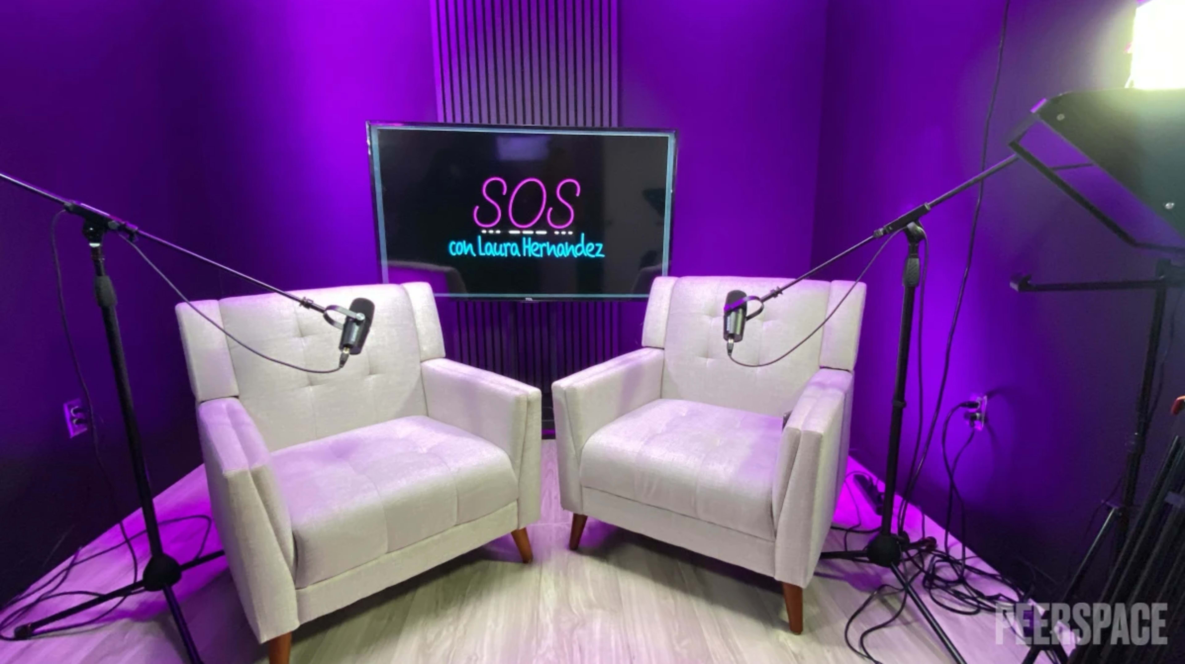 The image shows a brightly lit recording studio featuring two light-colored chairs, a large screen displaying the text "SOS con Laura Hernández," and microphones positioned for an interview setup.