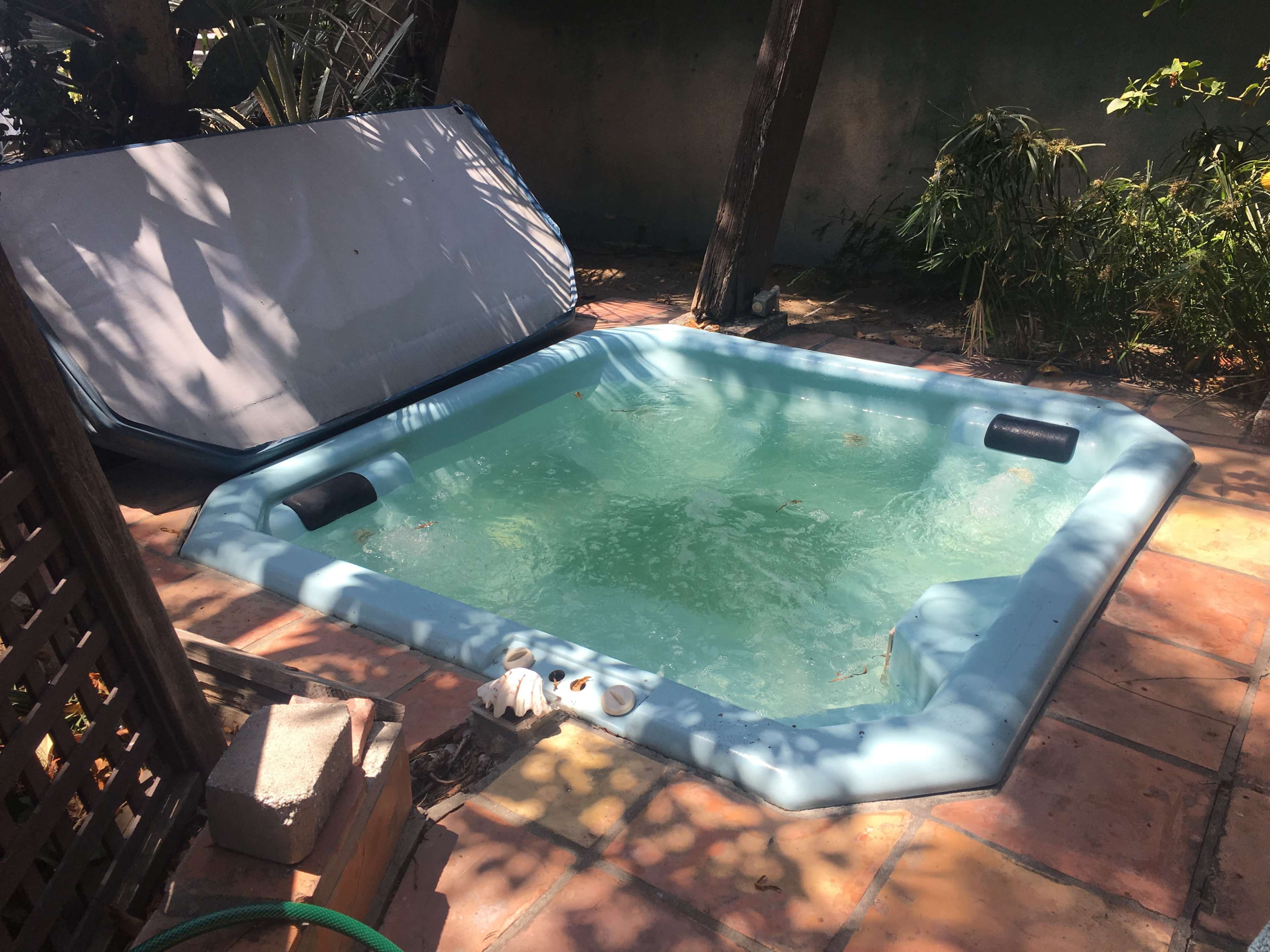 A rectangular hot tub filled with water sits in a shaded outdoor area surrounded by plants and tiled flooring.