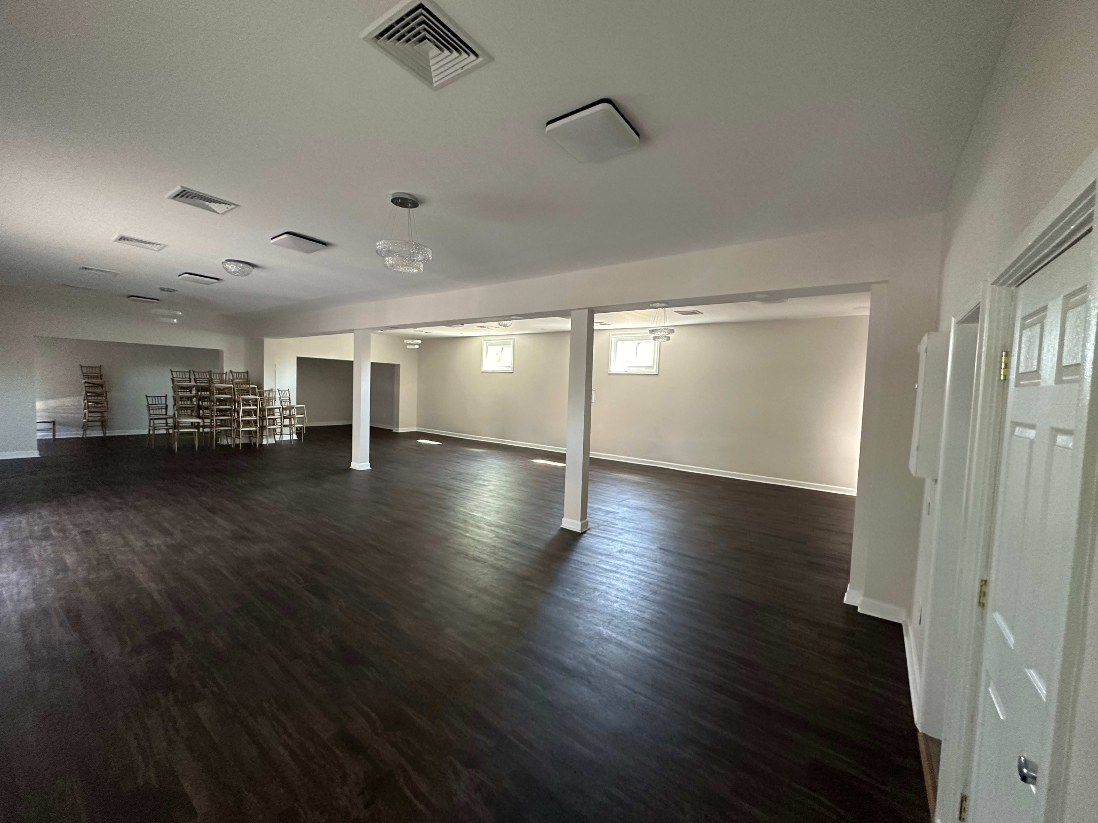 A spacious, empty room with dark wood flooring, several square columns, and stacks of chairs against the wall.