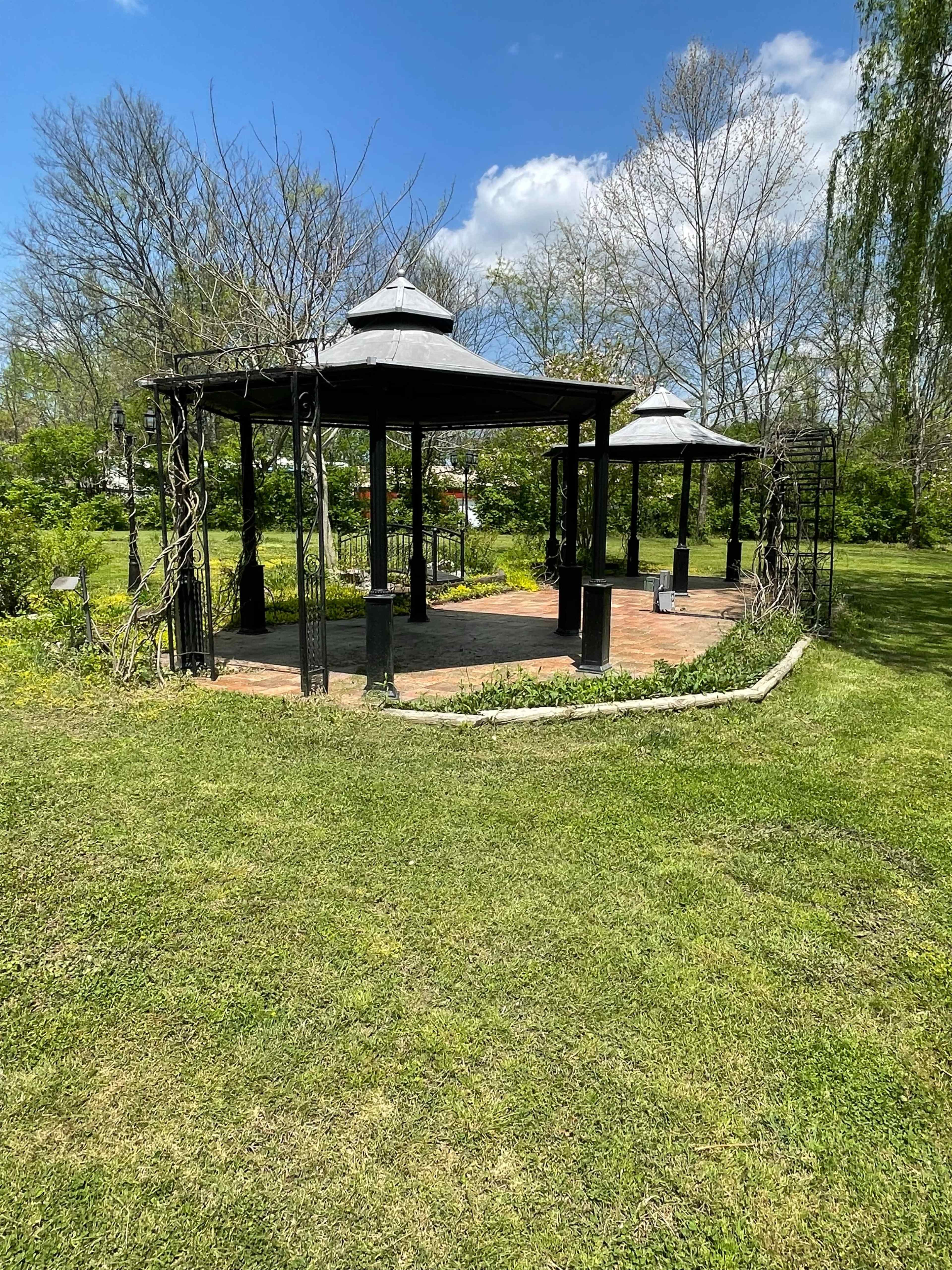 A pair of gazebo structures in a grassy area surrounded by trees and blue skies.