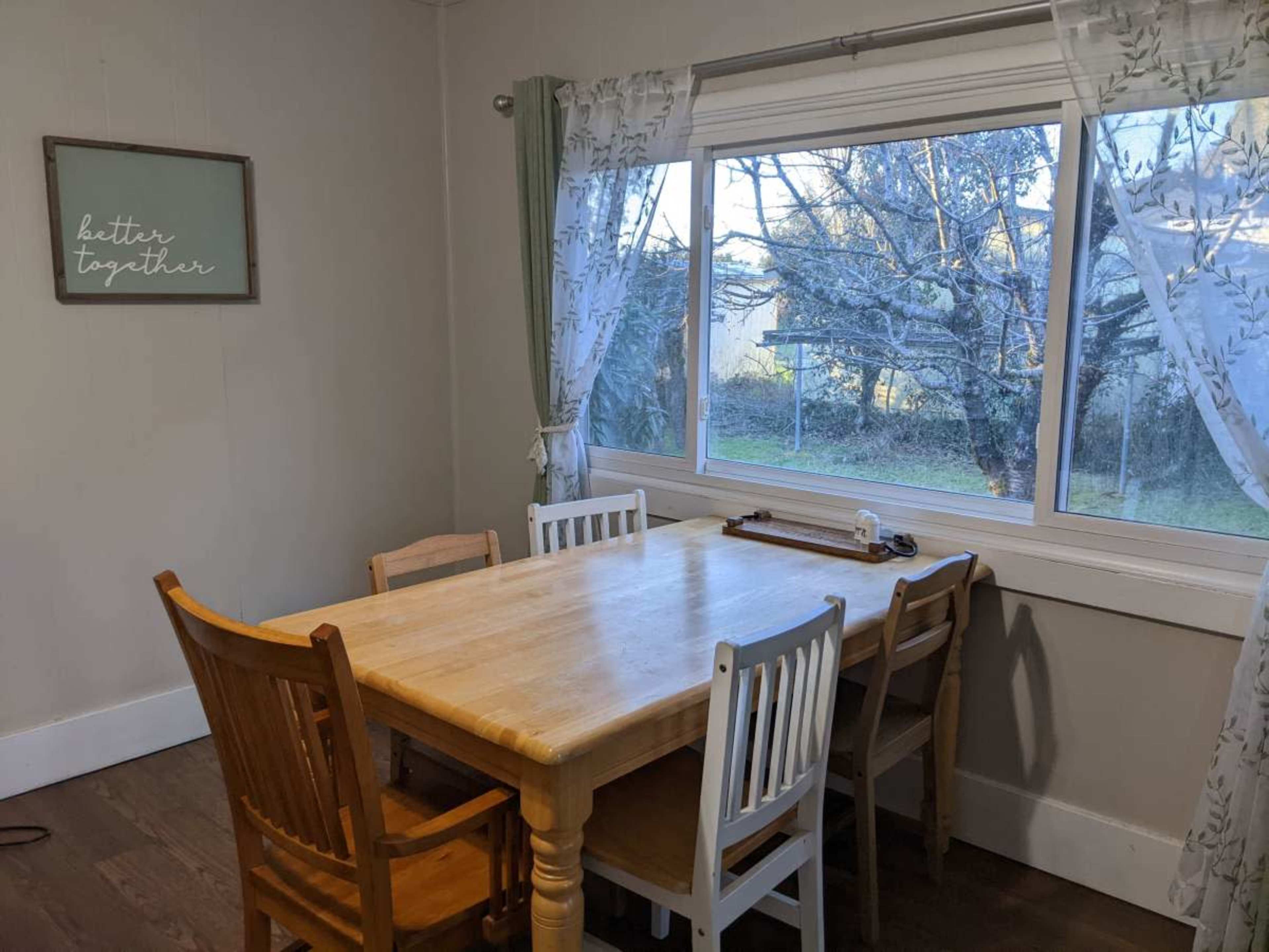 A wooden dining table with six chairs is positioned in a well-lit room next to a large window overlooking a garden.