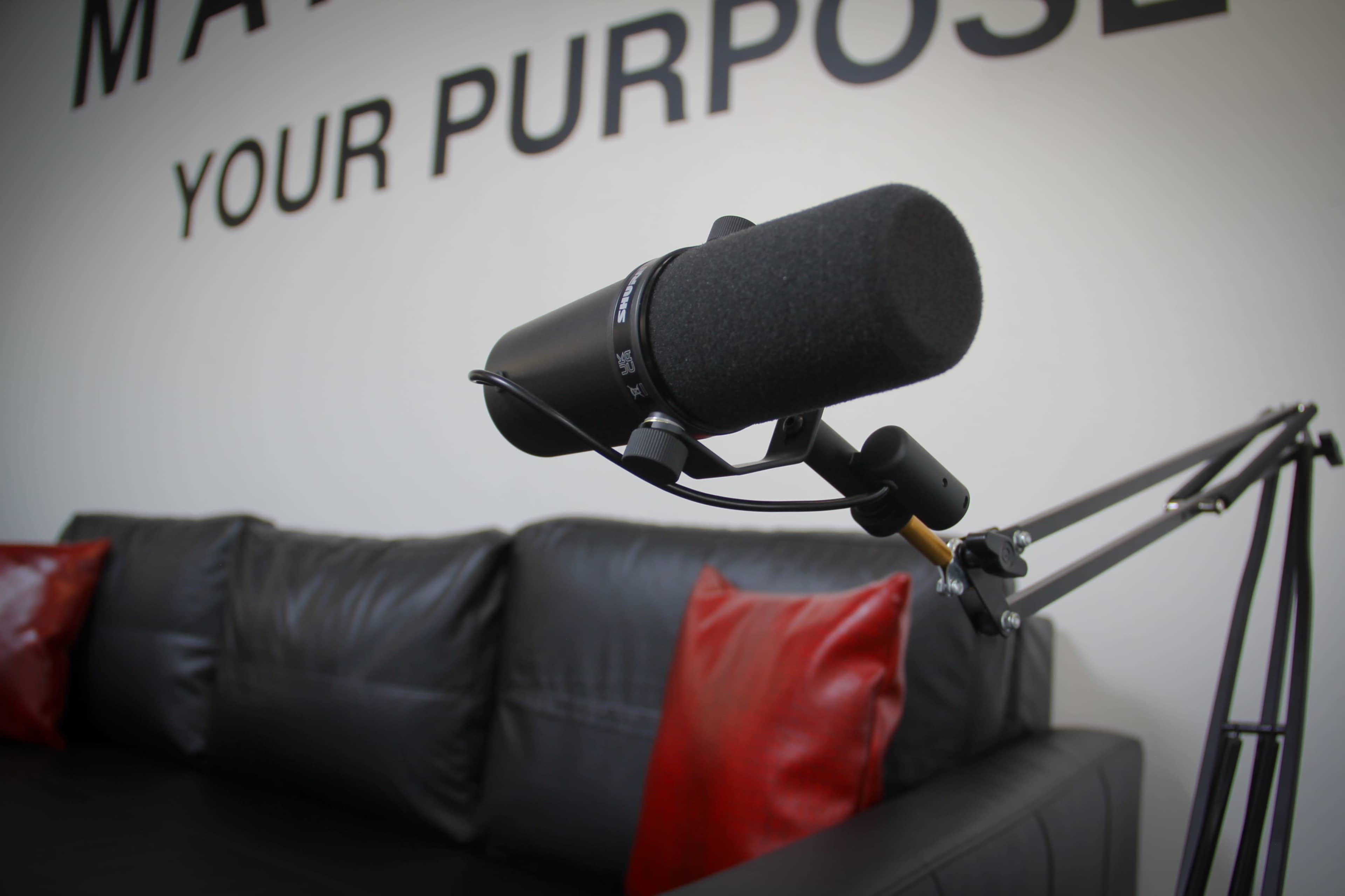 A microphone is mounted on a boom arm in front of a black couch with red pillows, against a wall featuring the words "MAKE YOUR PURPOSE."