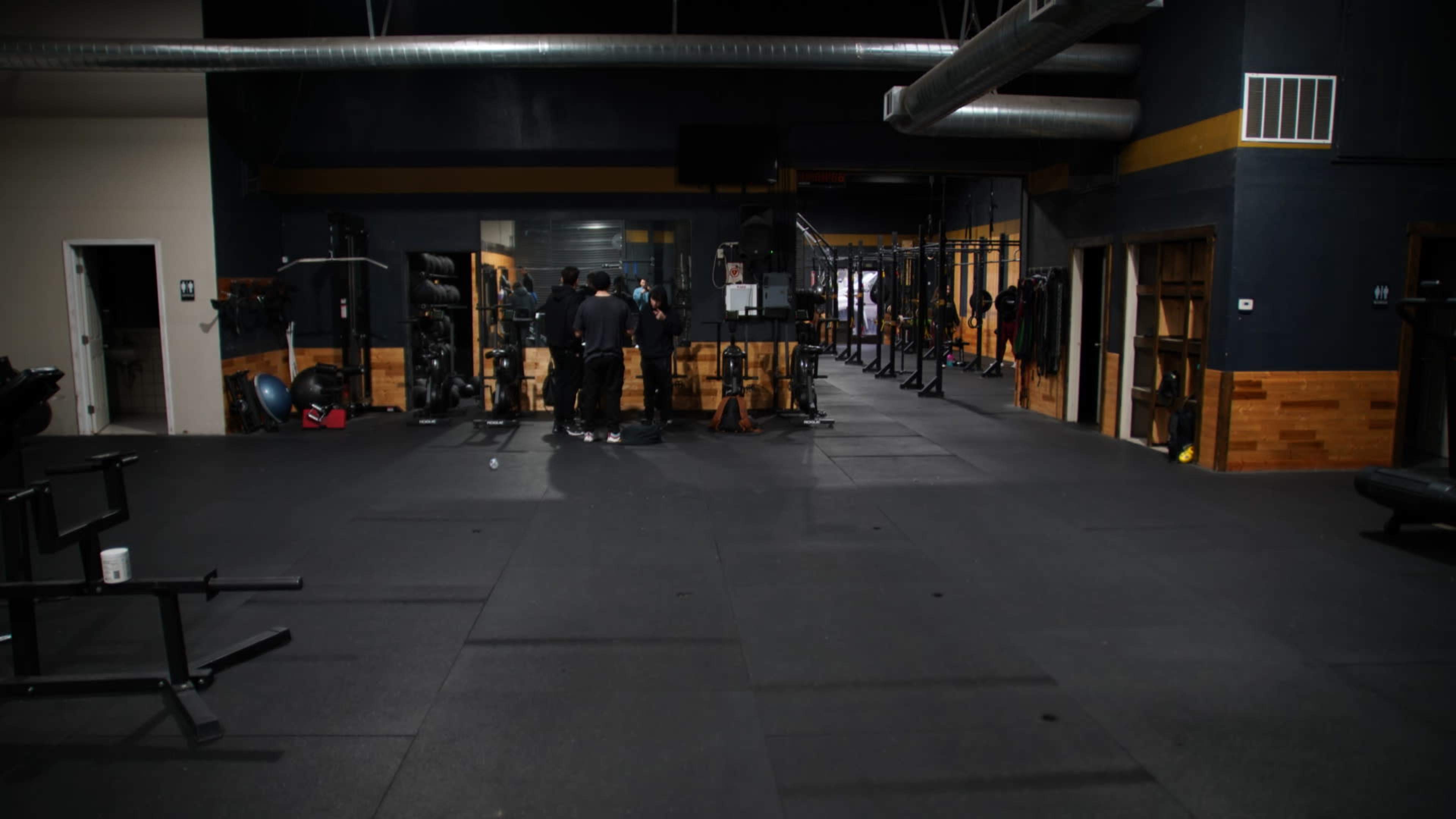 A gym interior features various workout equipment and several individuals gathered near the center.