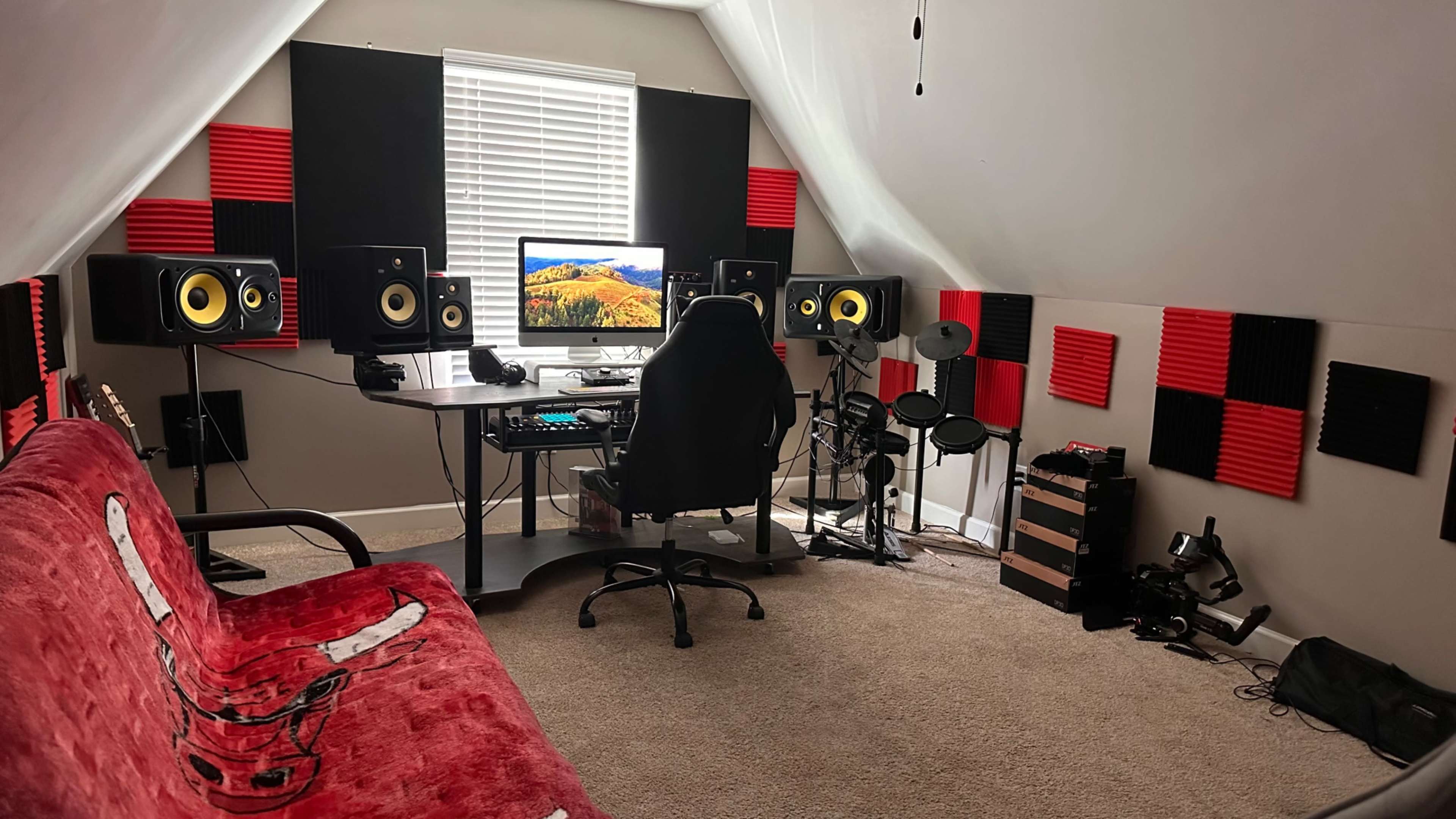 A room with acoustic panels on the walls contains a desk setup with a computer and speakers, a gaming chair, a red couch, and a small musical drum kit.