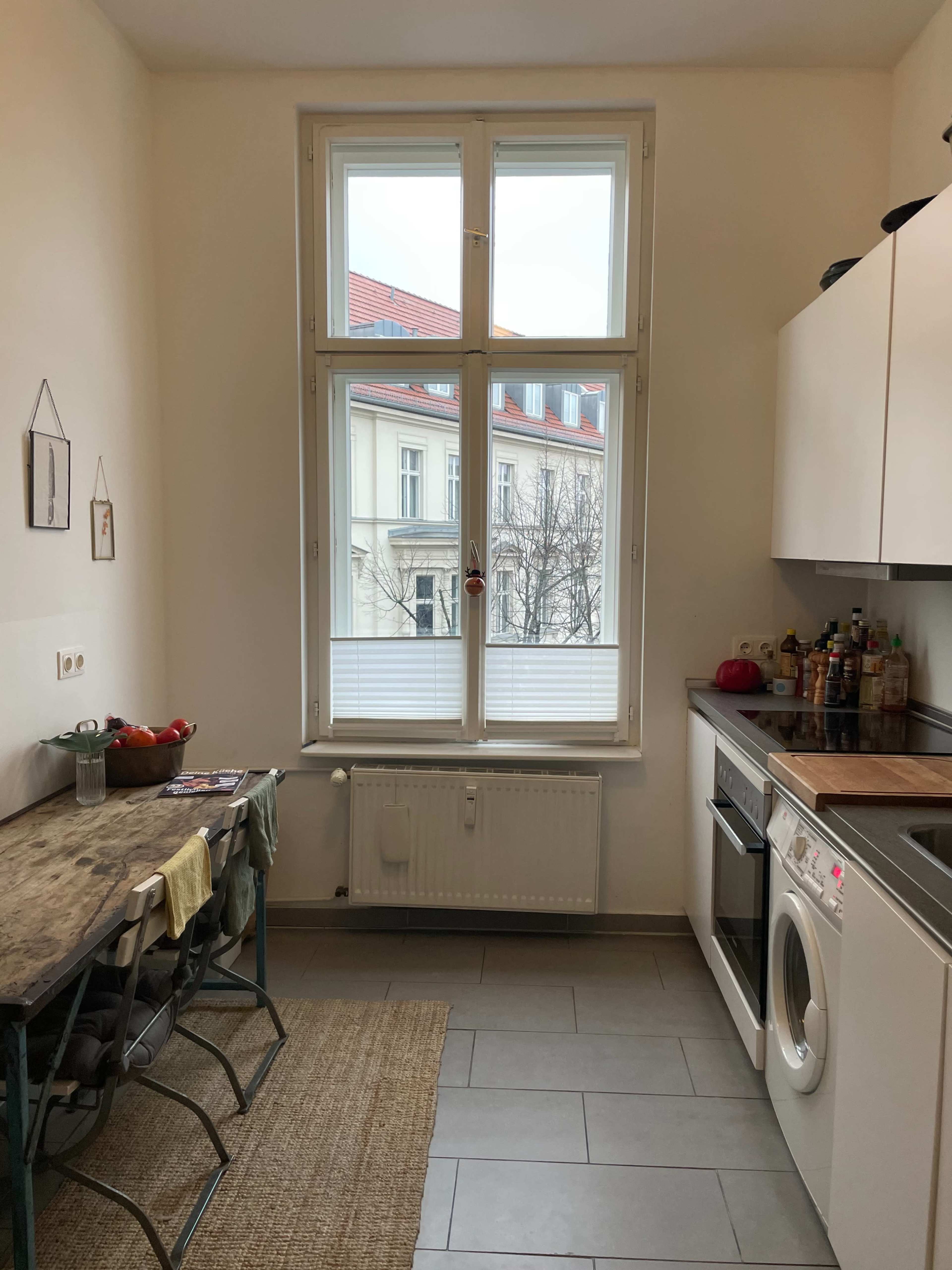 A small kitchen features a wooden table with two chairs, a washing machine, and a large window overlooking a building exterior.
