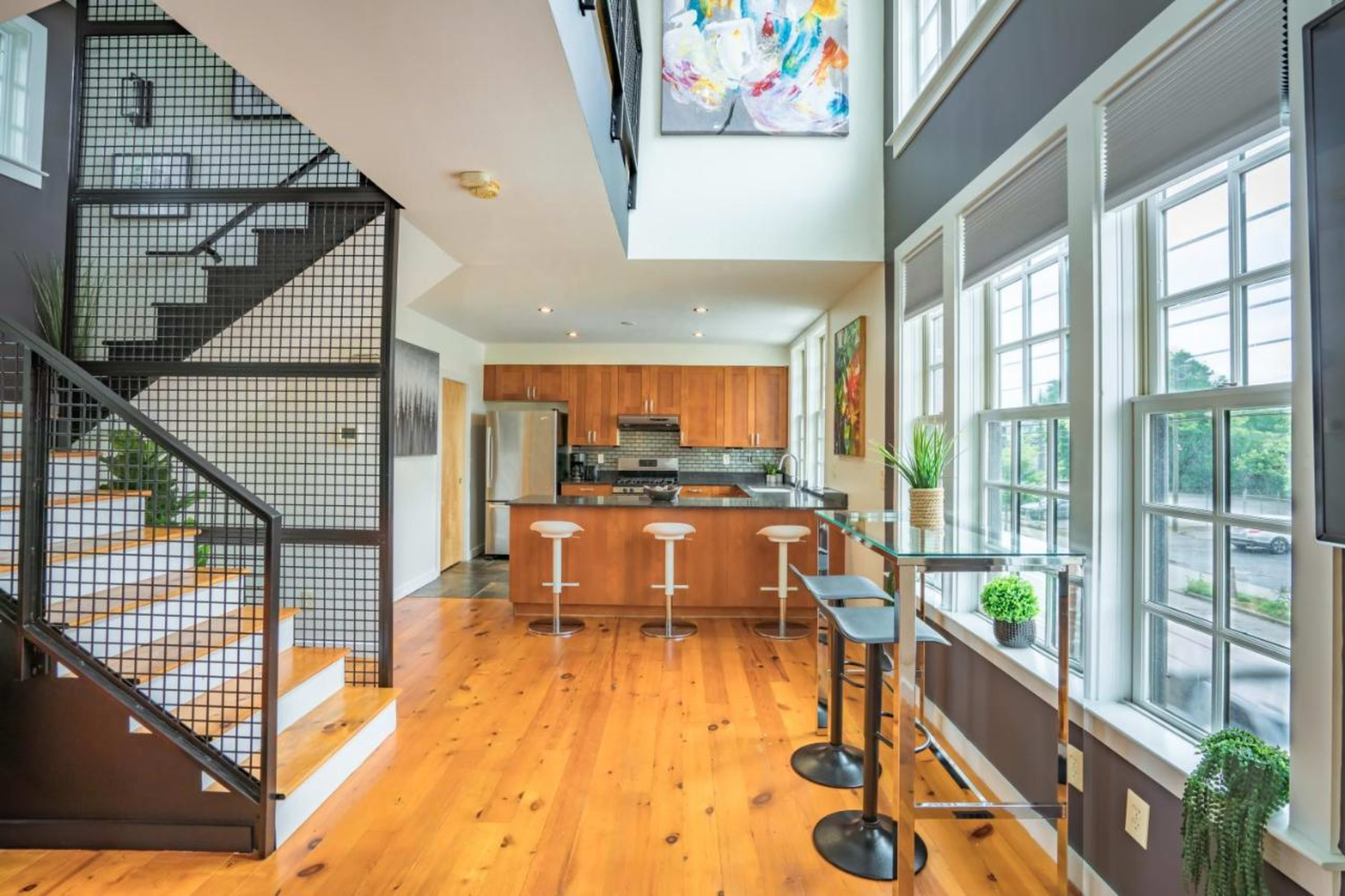 The image shows a modern kitchen and dining area with hardwood floors, large windows, and a staircase featuring a metal railing.