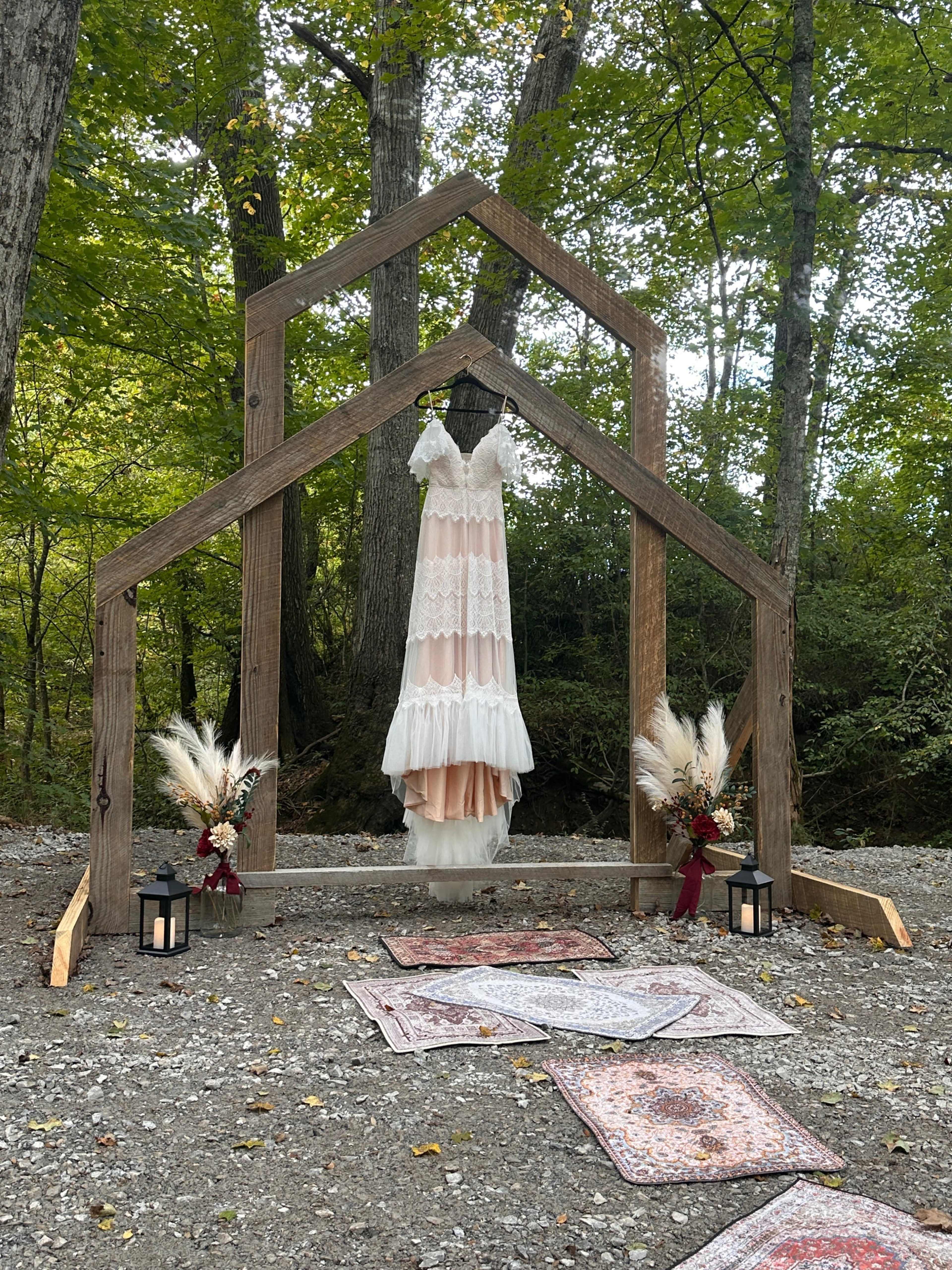 A white lace dress hangs from a wooden arch in a forested area, surrounded by decorative plants and lanterns on a pathway of patterned rugs.