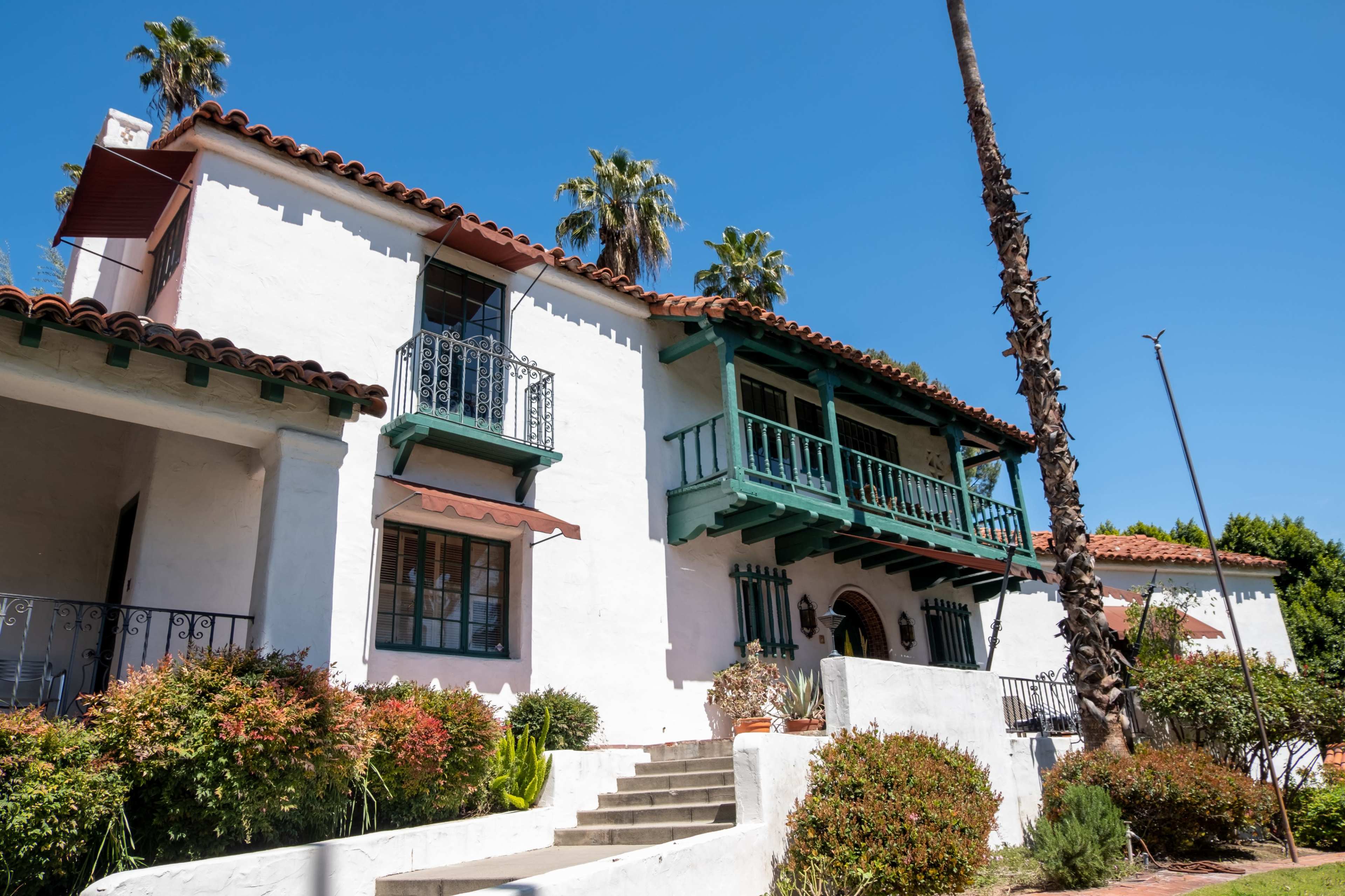 The image shows a two-story white Mediterranean-style house with green balconies, surrounded by palm trees and landscaped shrubs.