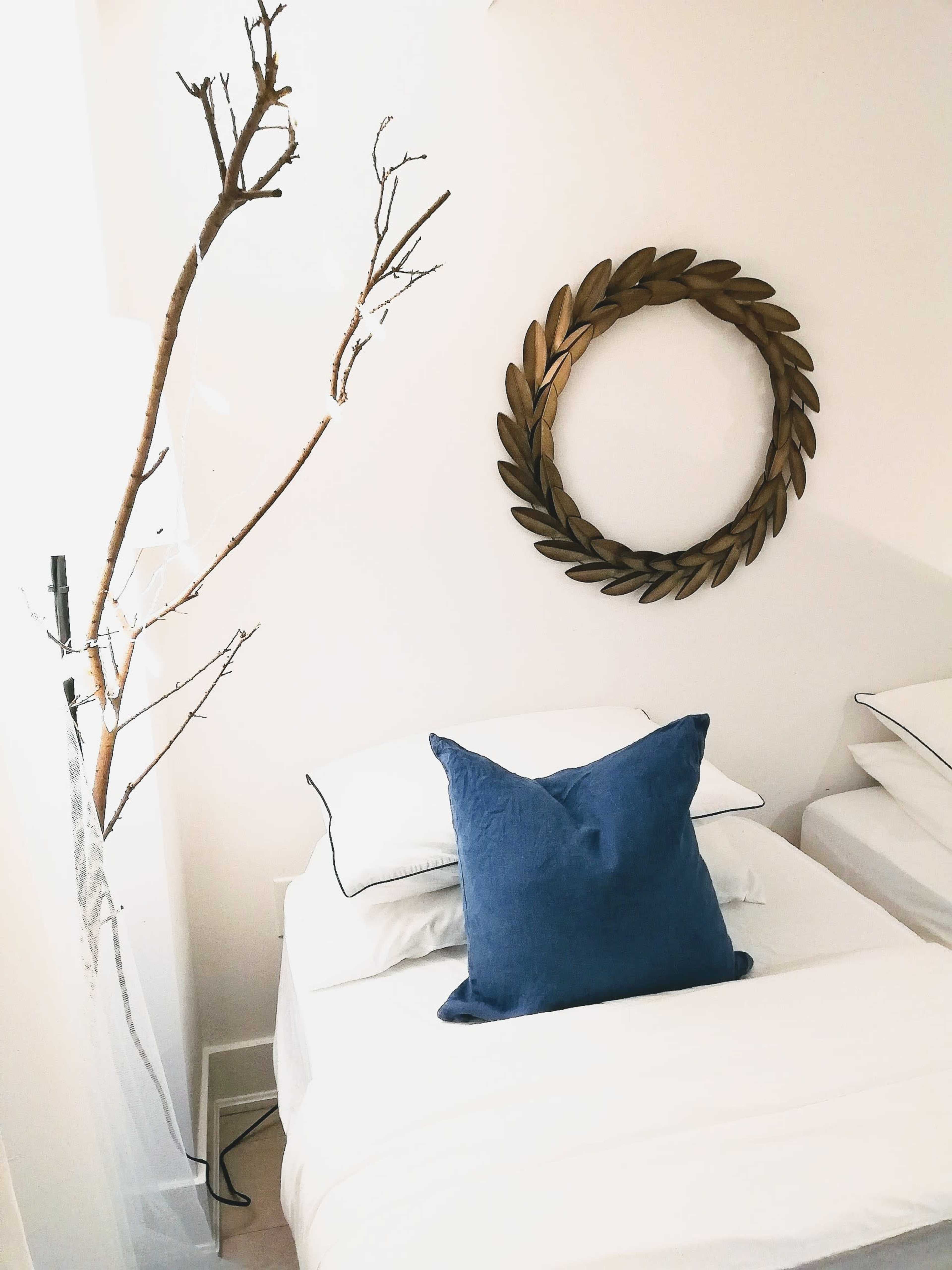 A bed with a blue pillow is positioned against a white wall featuring a decorative circular wall piece and a bare tree branch in the corner.