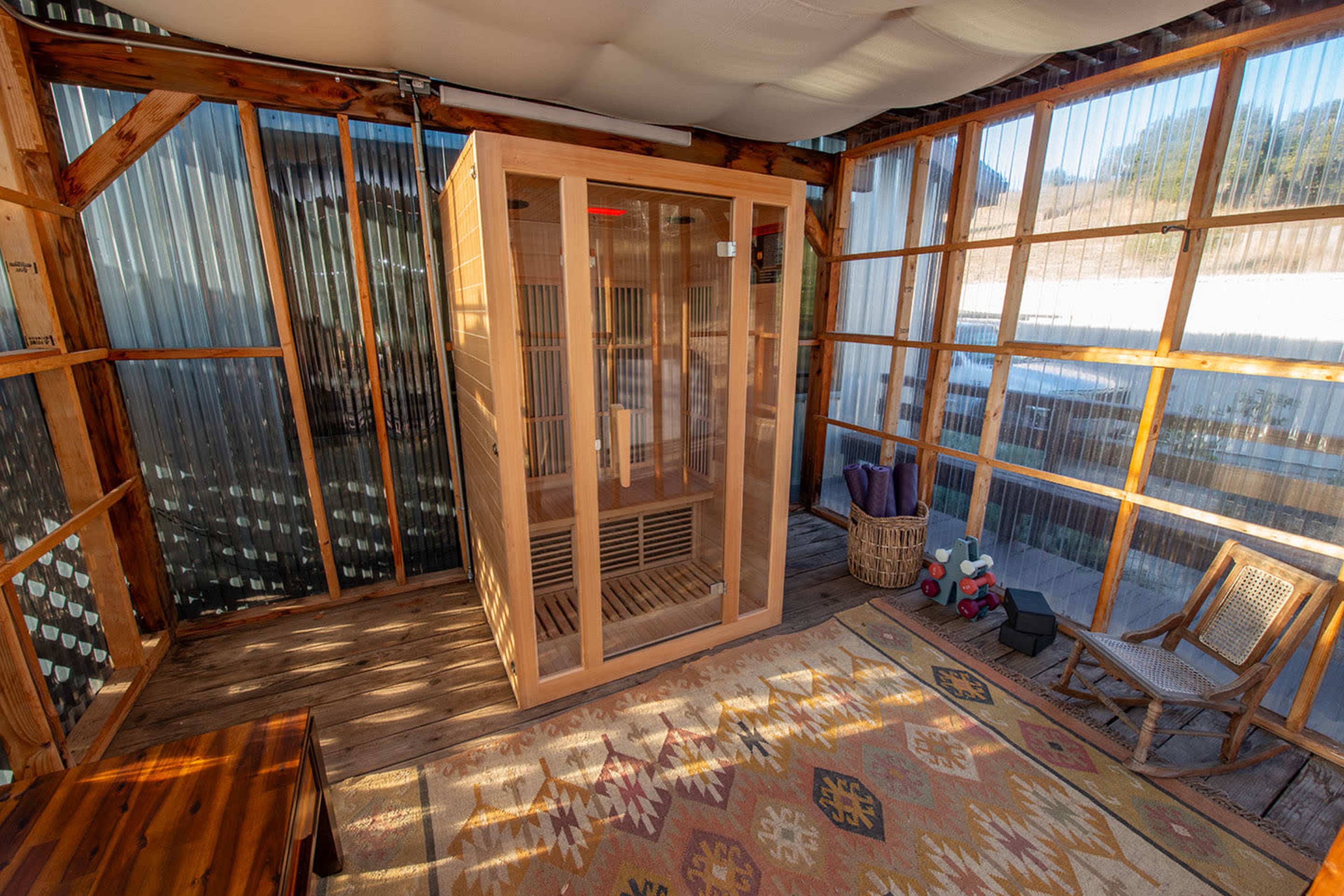 The image shows a wooden sauna cabin placed in a room with glass walls, surrounded by wooden furniture and an area rug.