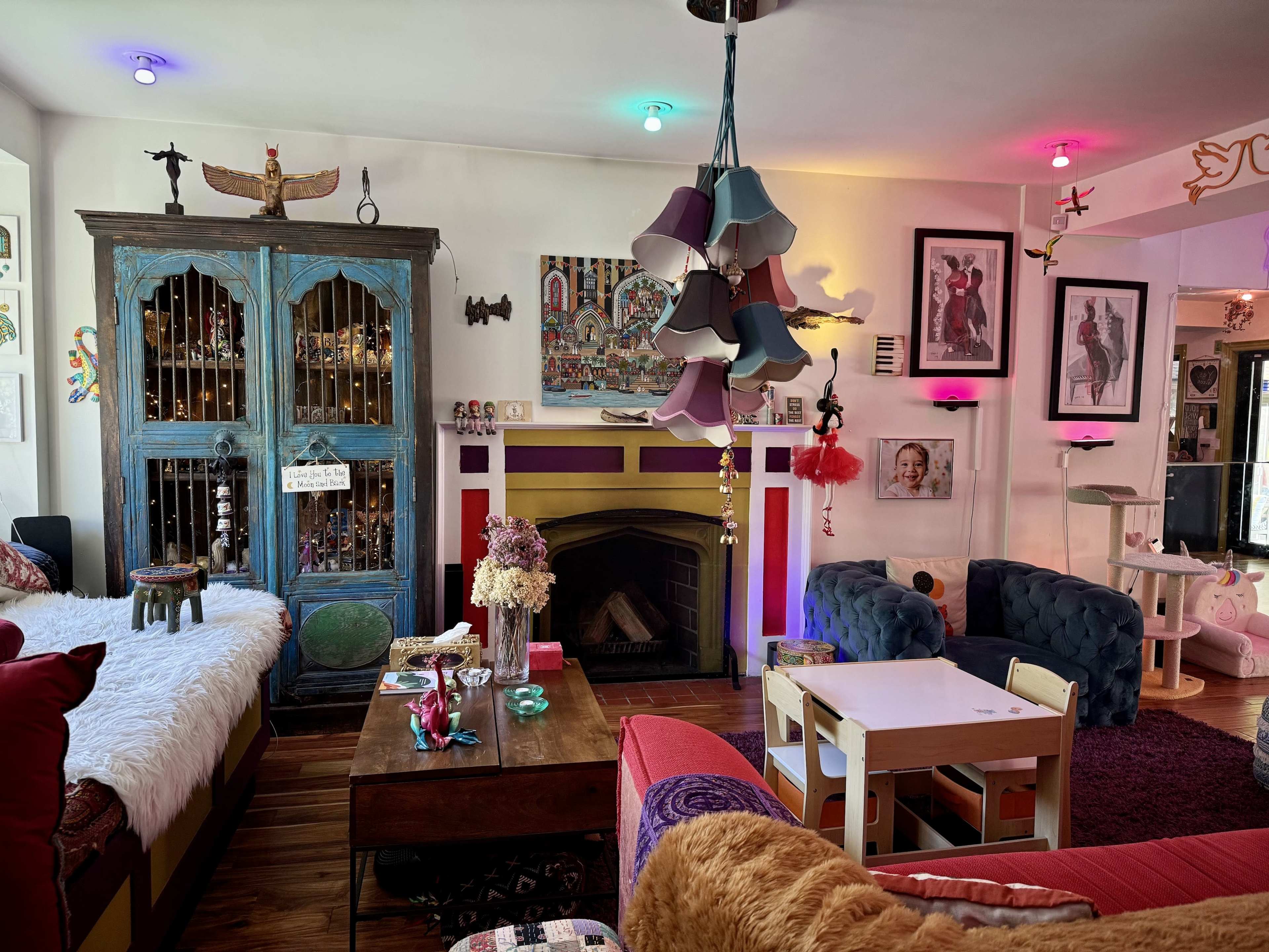 The image shows a colorful living room with eclectic decor, featuring a blue cabinet, a fireplace, various artworks on the walls, and playful furniture.