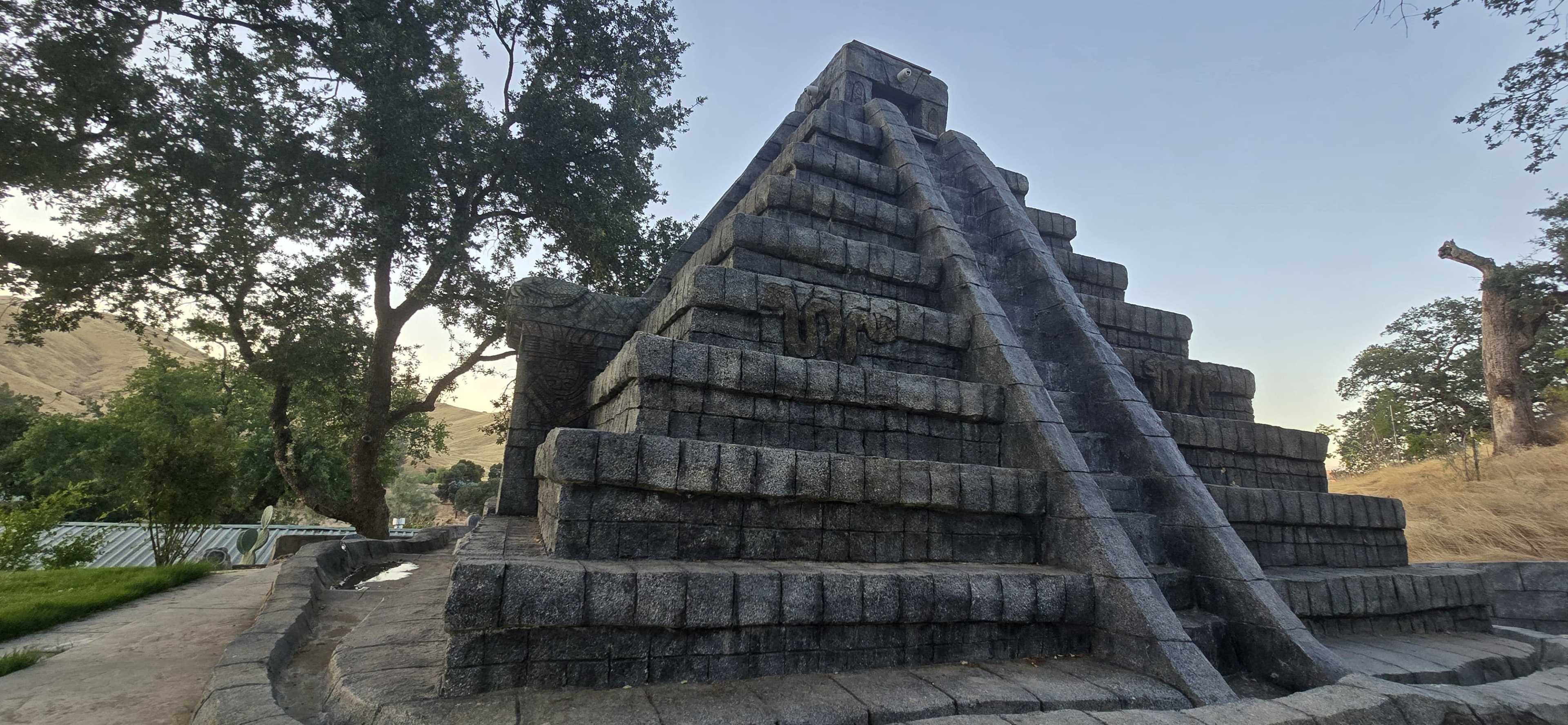The image shows a pyramid-shaped stone structure with steps leading up one side, surrounded by trees and open land.