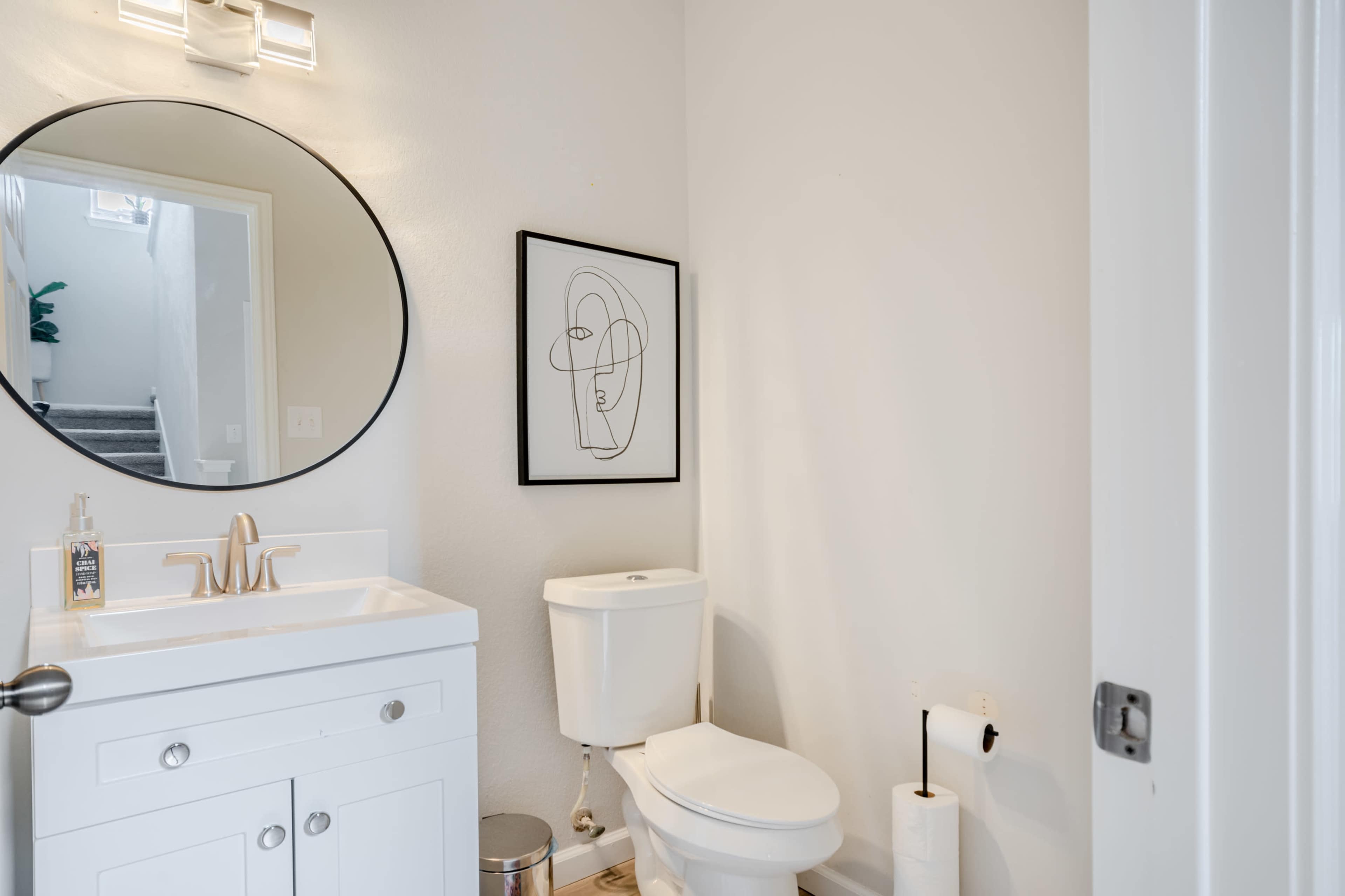A small bathroom featuring a circular mirror above a white vanity with a sink, a toilet, and a framed abstract artwork on the wall.