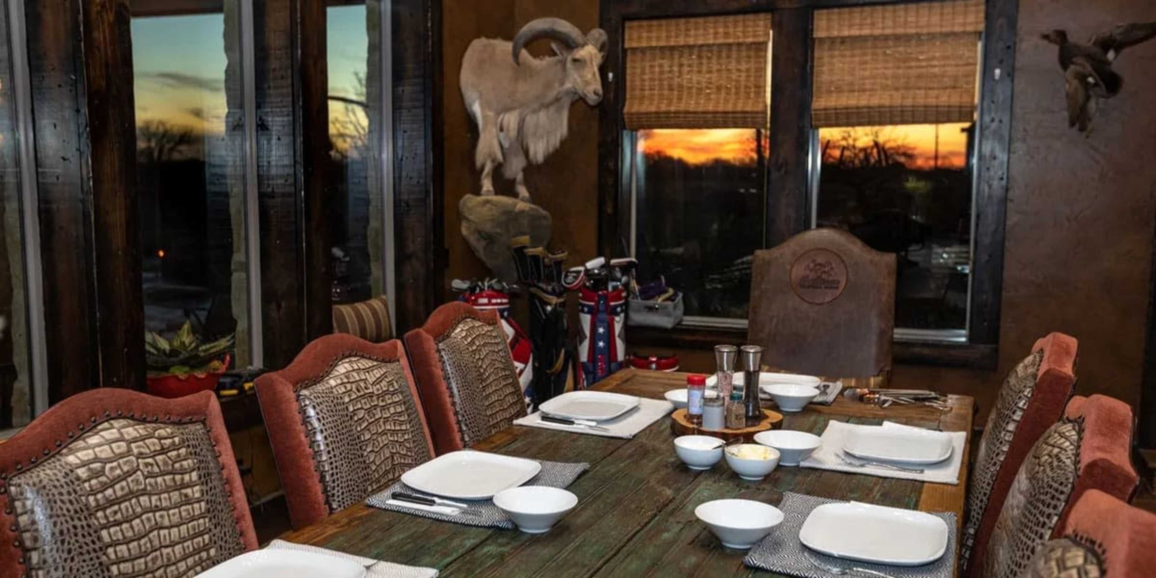 The image shows a dining table set with plates and bowls in a rustic room featuring a sunset view and mounted animal trophies on the walls.