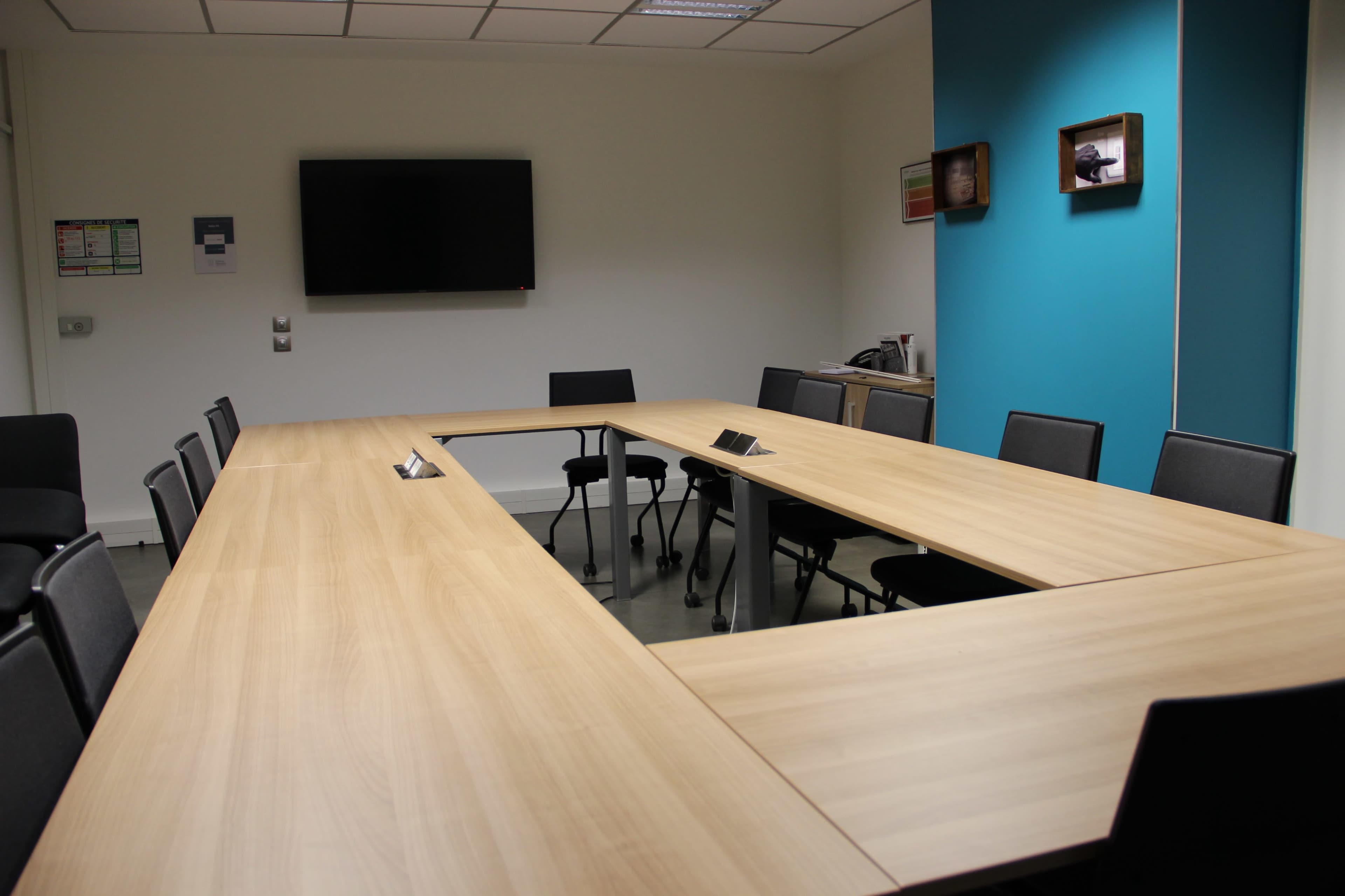 A modern conference room features a long wooden table surrounded by black chairs, with a wall-mounted television and blue accent wall.