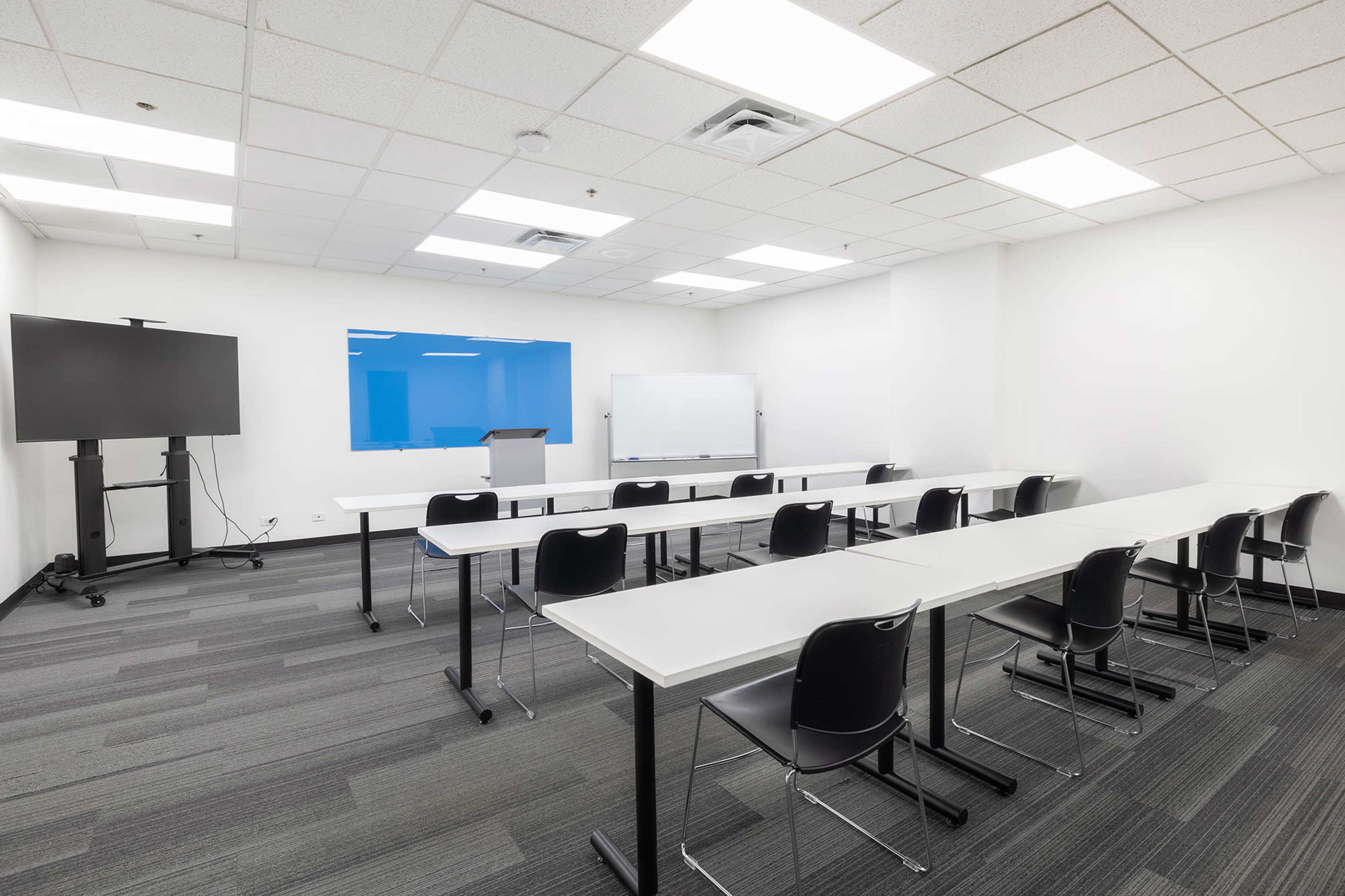 The image shows a modern classroom with several rows of tables and chairs, a large screen, a podium, and a whiteboard.