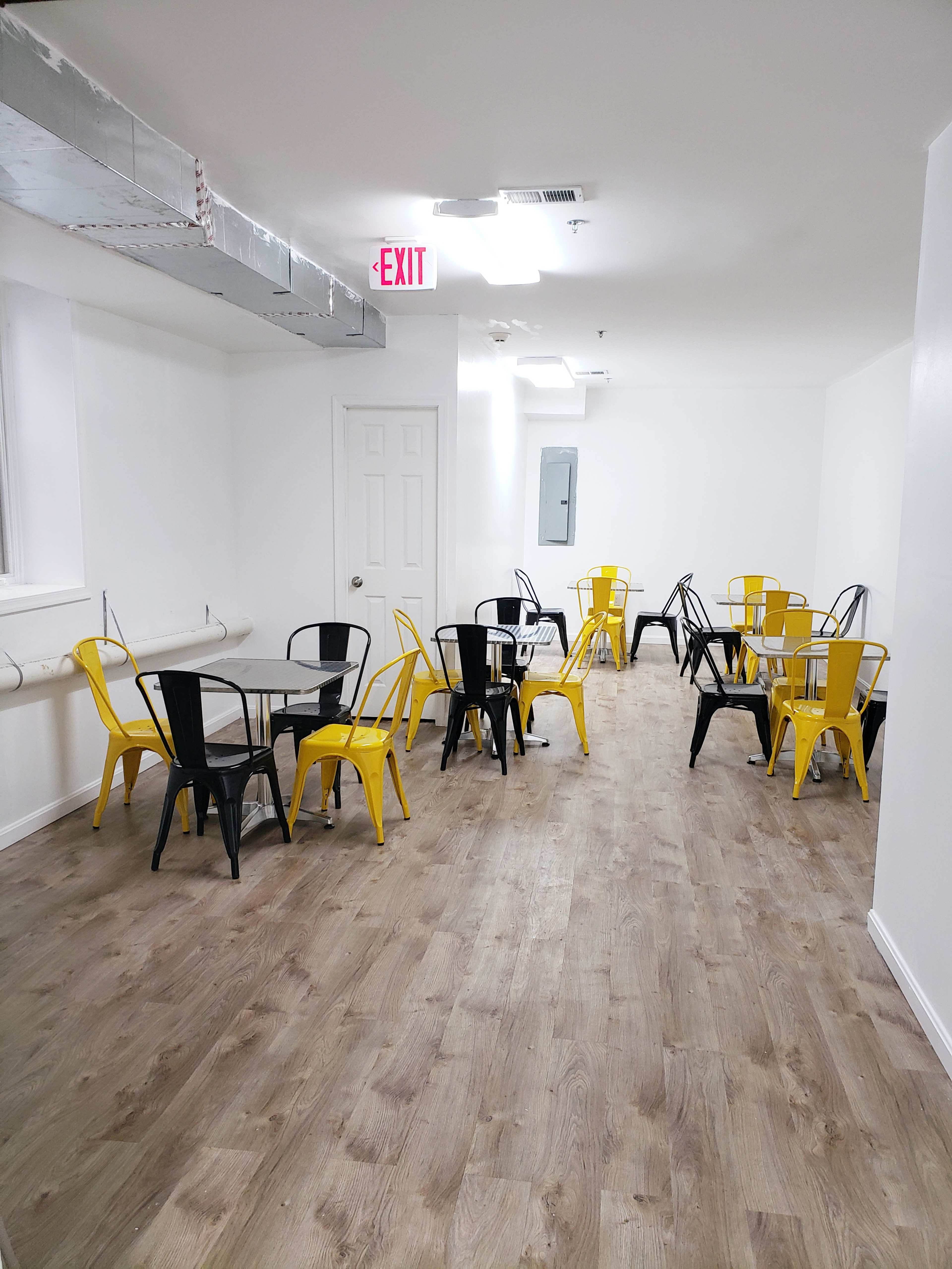 A spacious, well-lit room with wooden flooring, featuring several metal tables with black and yellow chairs arranged throughout.