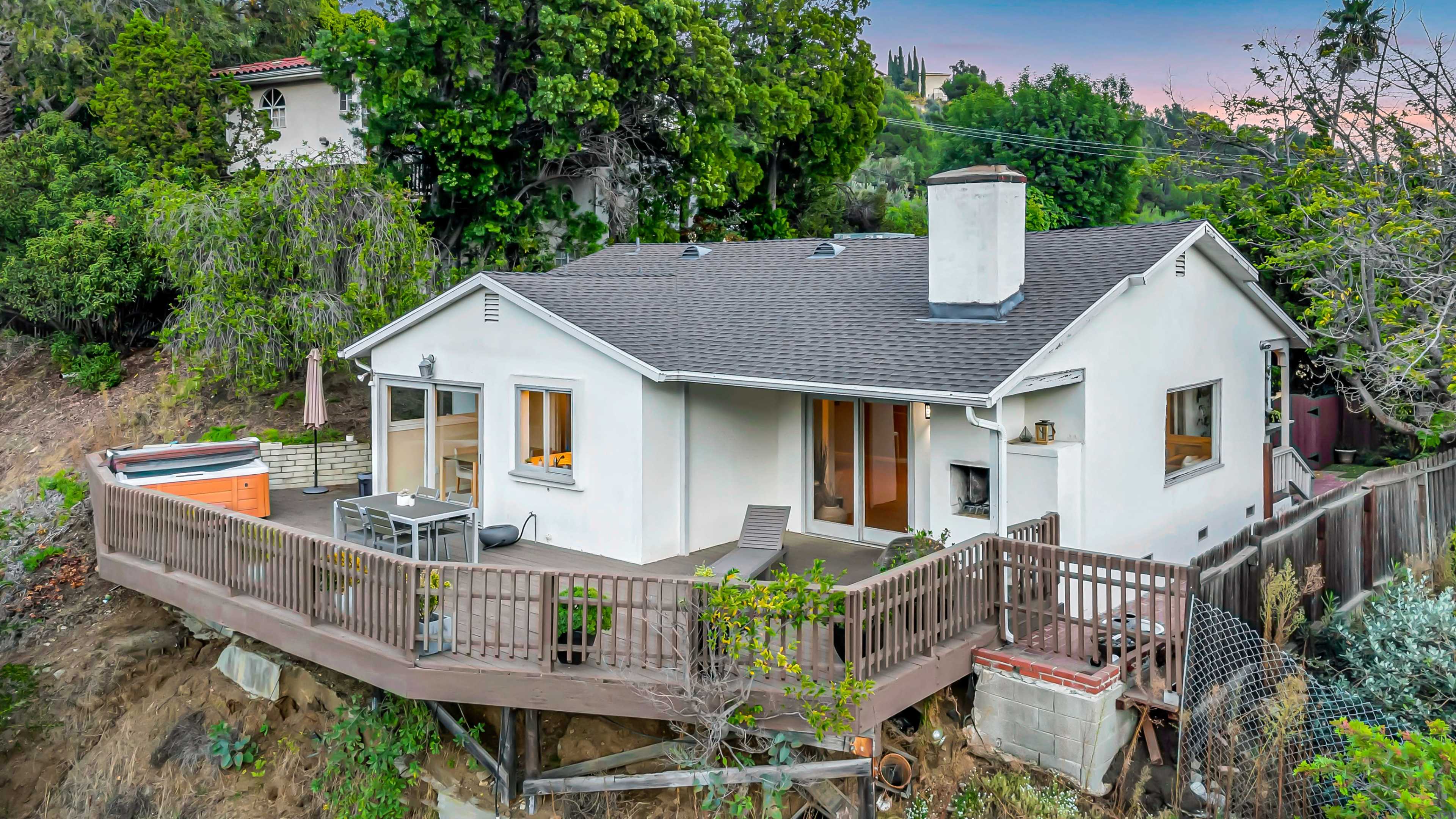 The image shows a single-story house with a wooden deck, surrounded by trees and located on a slope.