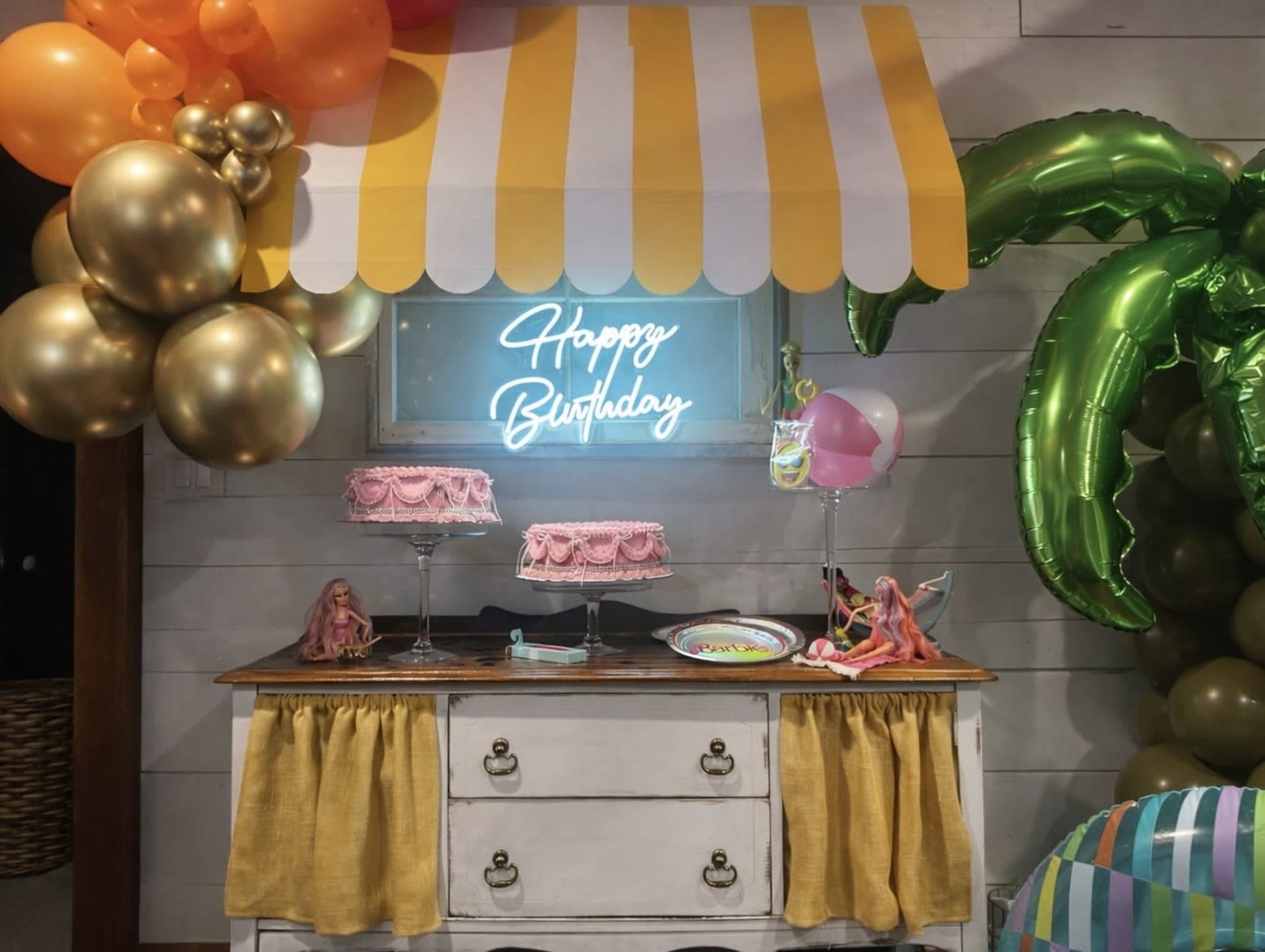 The image features a birthday celebration setup with a decorated table displaying pink cakes, a neon "Happy Birthday" sign, and vibrant balloons.