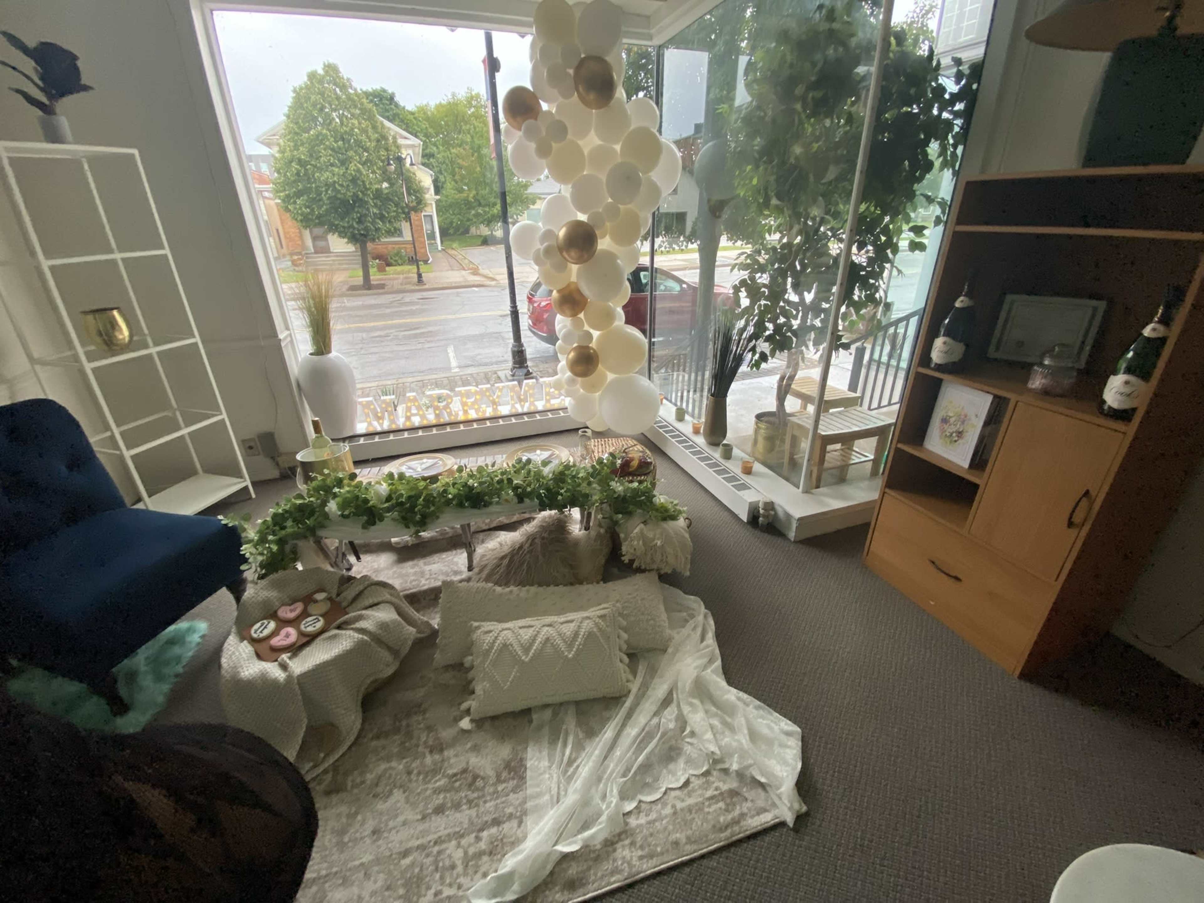 A cozy indoor setup with a rug, various cushions, and a coffee table adorned with decorative items, surrounded by large windows and greenery outside.