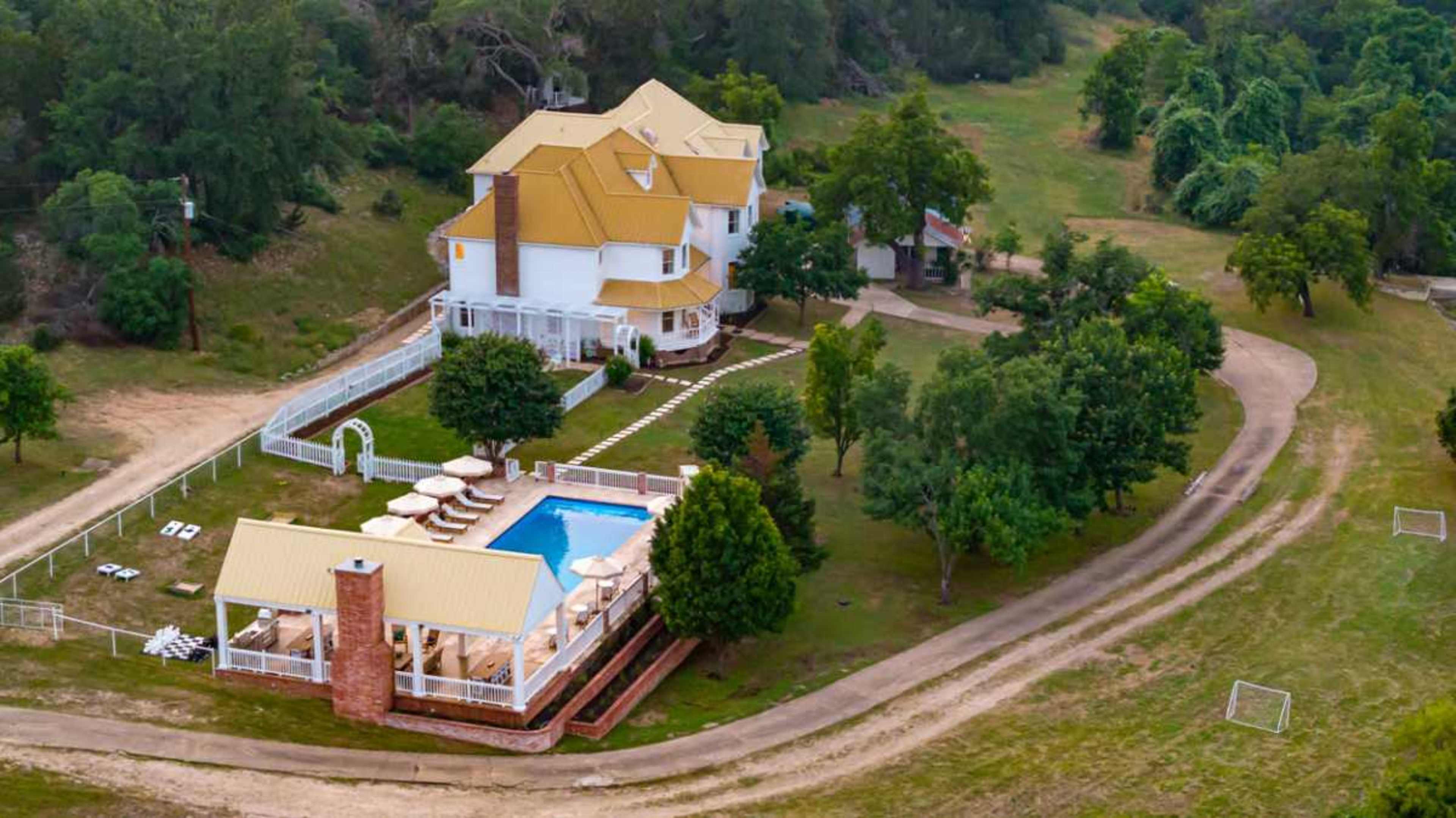 The image shows a large, two-story house with a yellow roof, surrounded by a green landscape, a swimming pool, and several lounge chairs.
