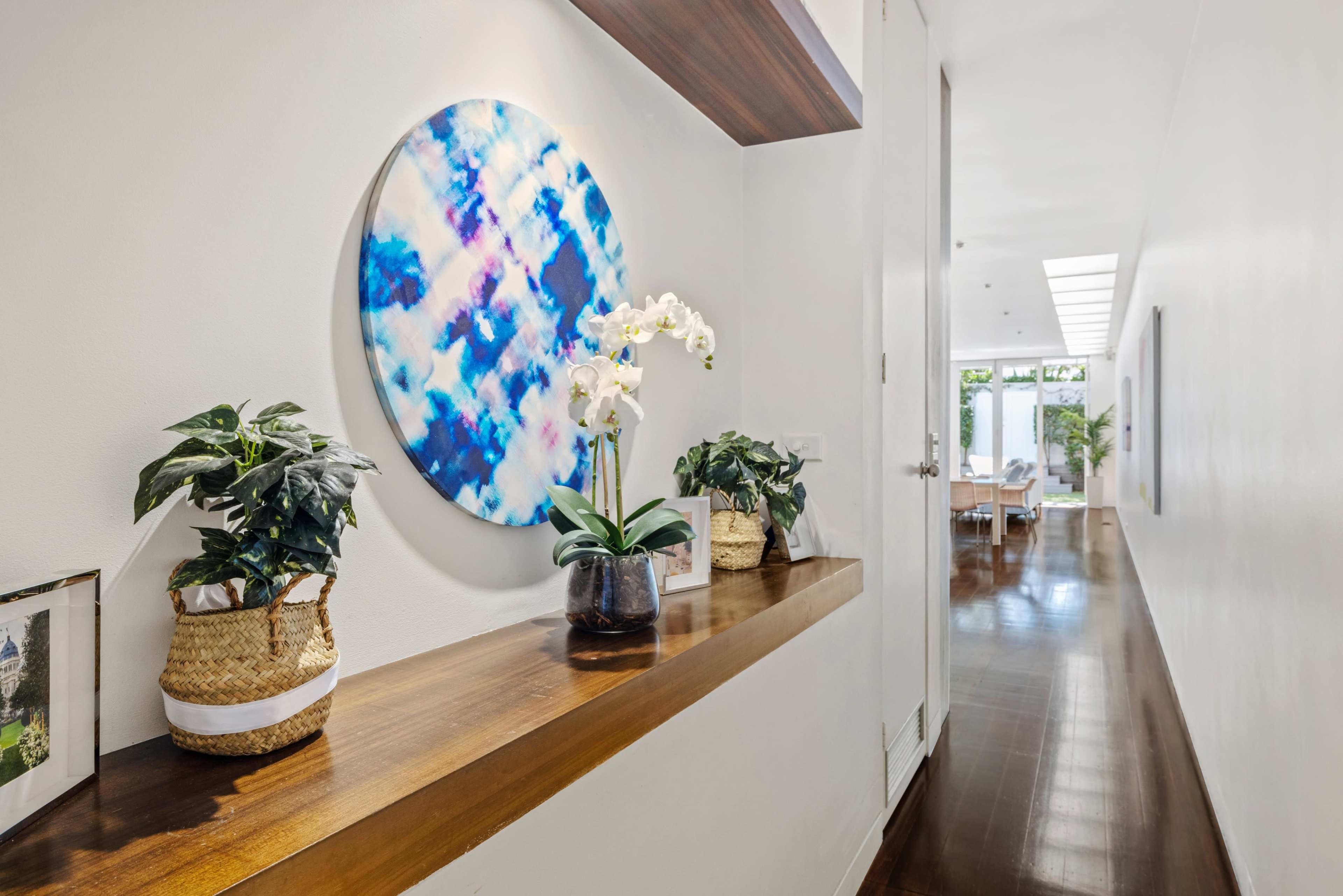 A narrow hallway features a wooden ledge adorned with potted plants and an abstract circular artwork, leading towards a bright room at the end.
