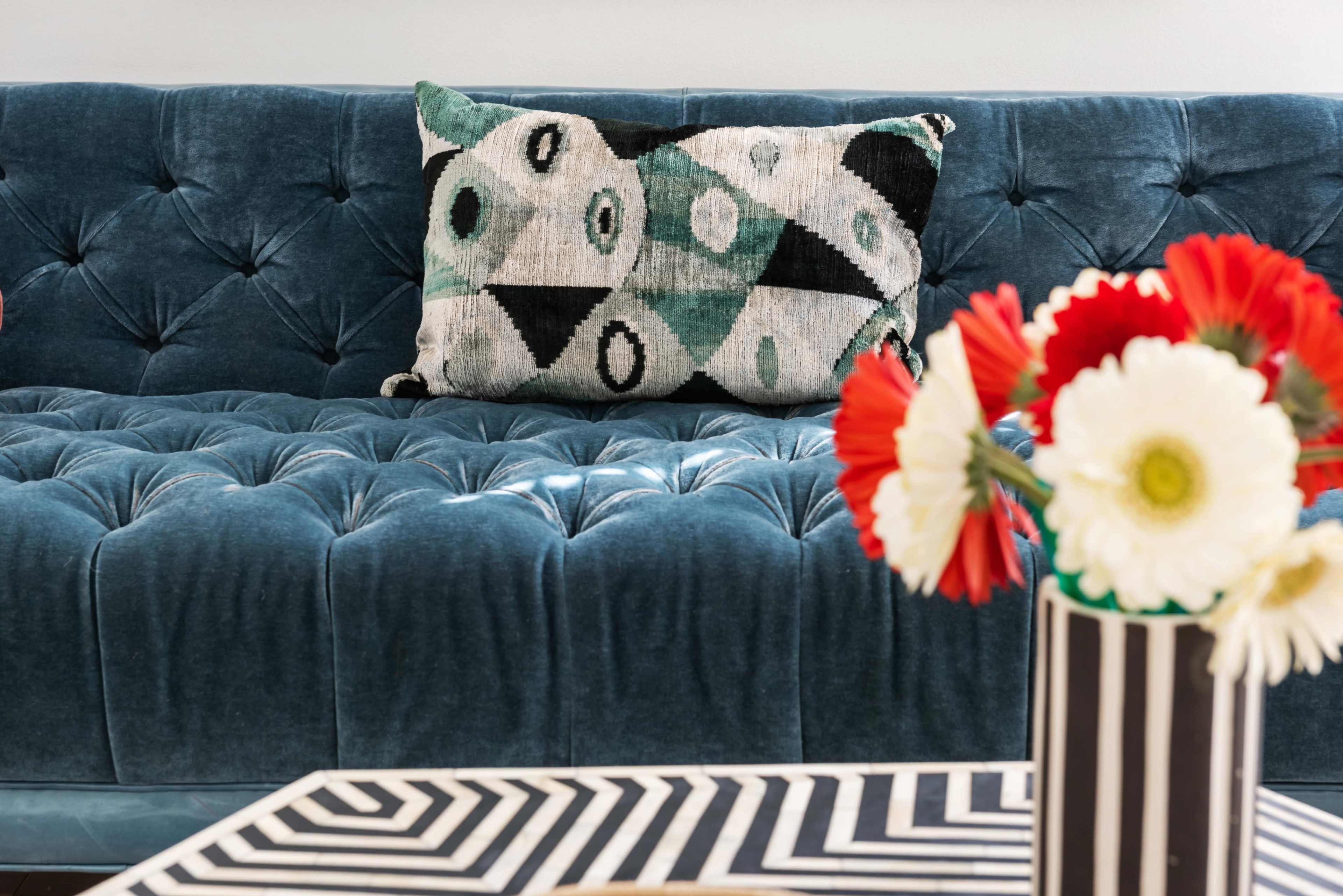 A tufted blue velvet sofa with a patterned pillow sits beside a striped vase filled with flowers on a geometric coffee table.