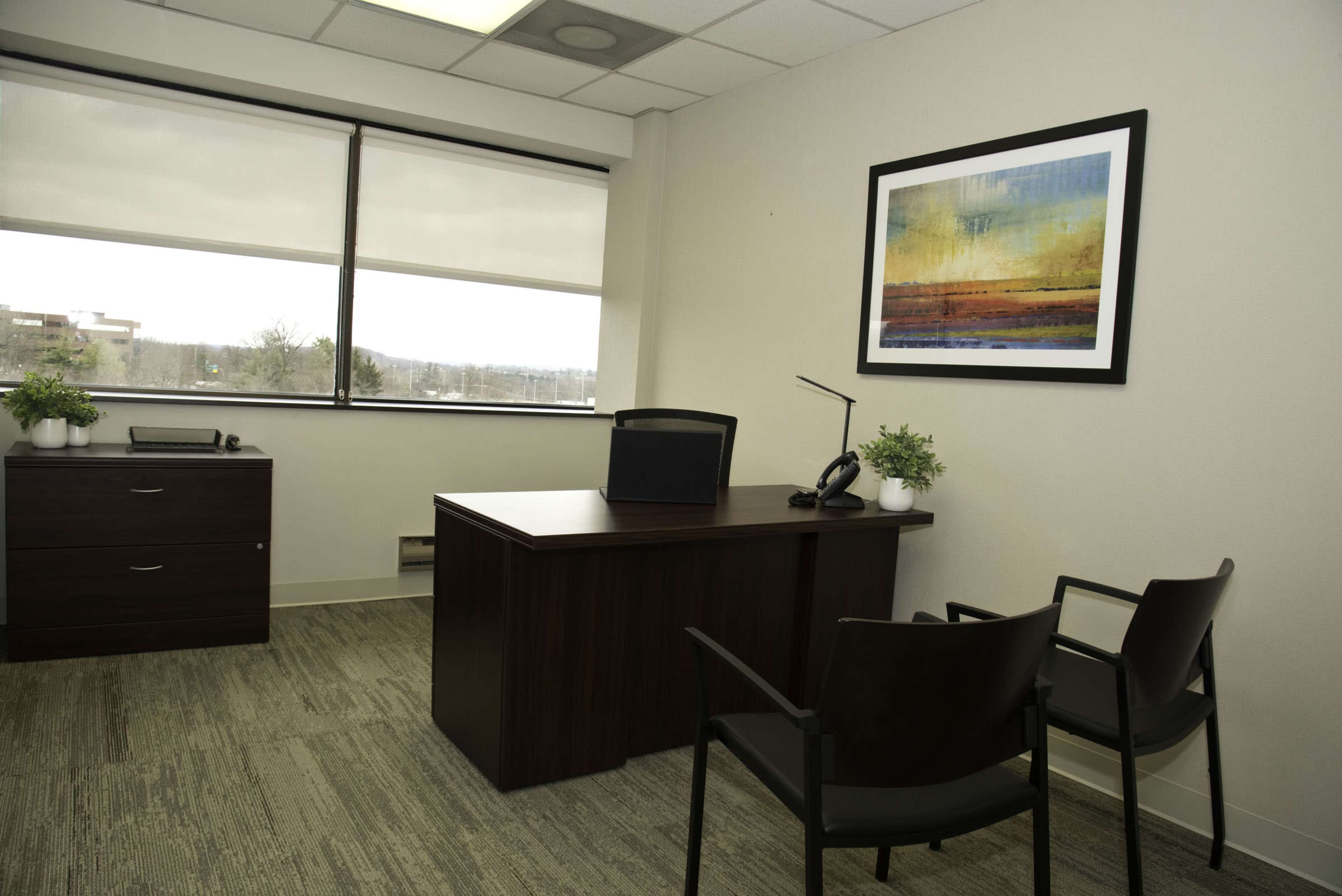 The image shows a minimalist office space featuring a wooden desk with a chair, a small filing cabinet, and a window with blinds.